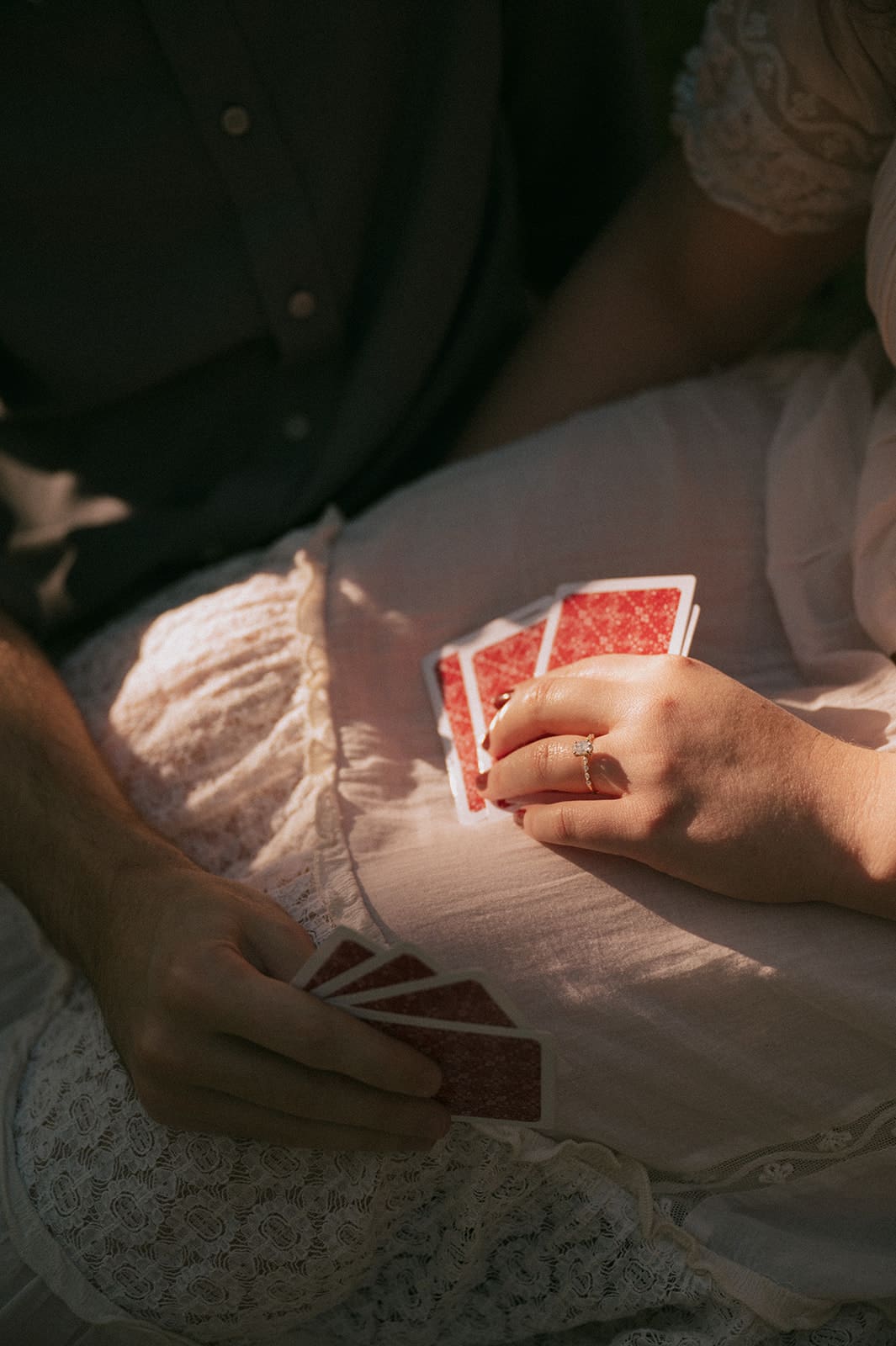 Two people sitting close together, holding playing cards with red backs, one person wearing a white lace dress.