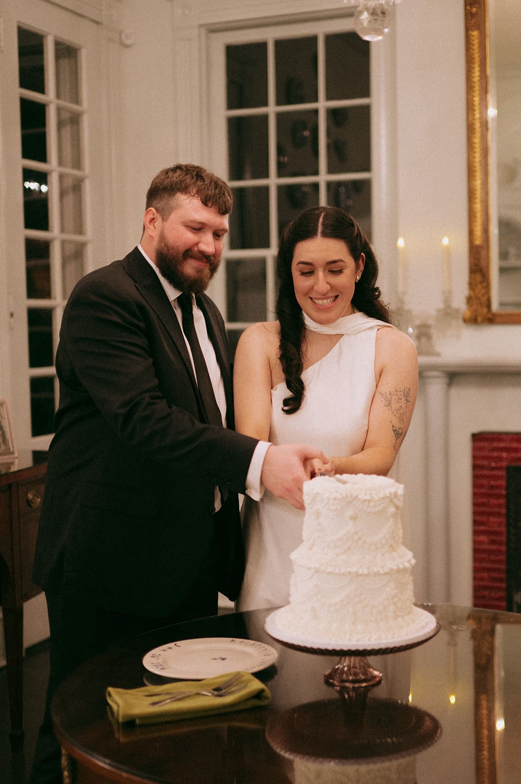 A couple in formal attire stands together cutting a white, three-tiered cake on a table in a room with white walls and large windows at Woodbine Mansion