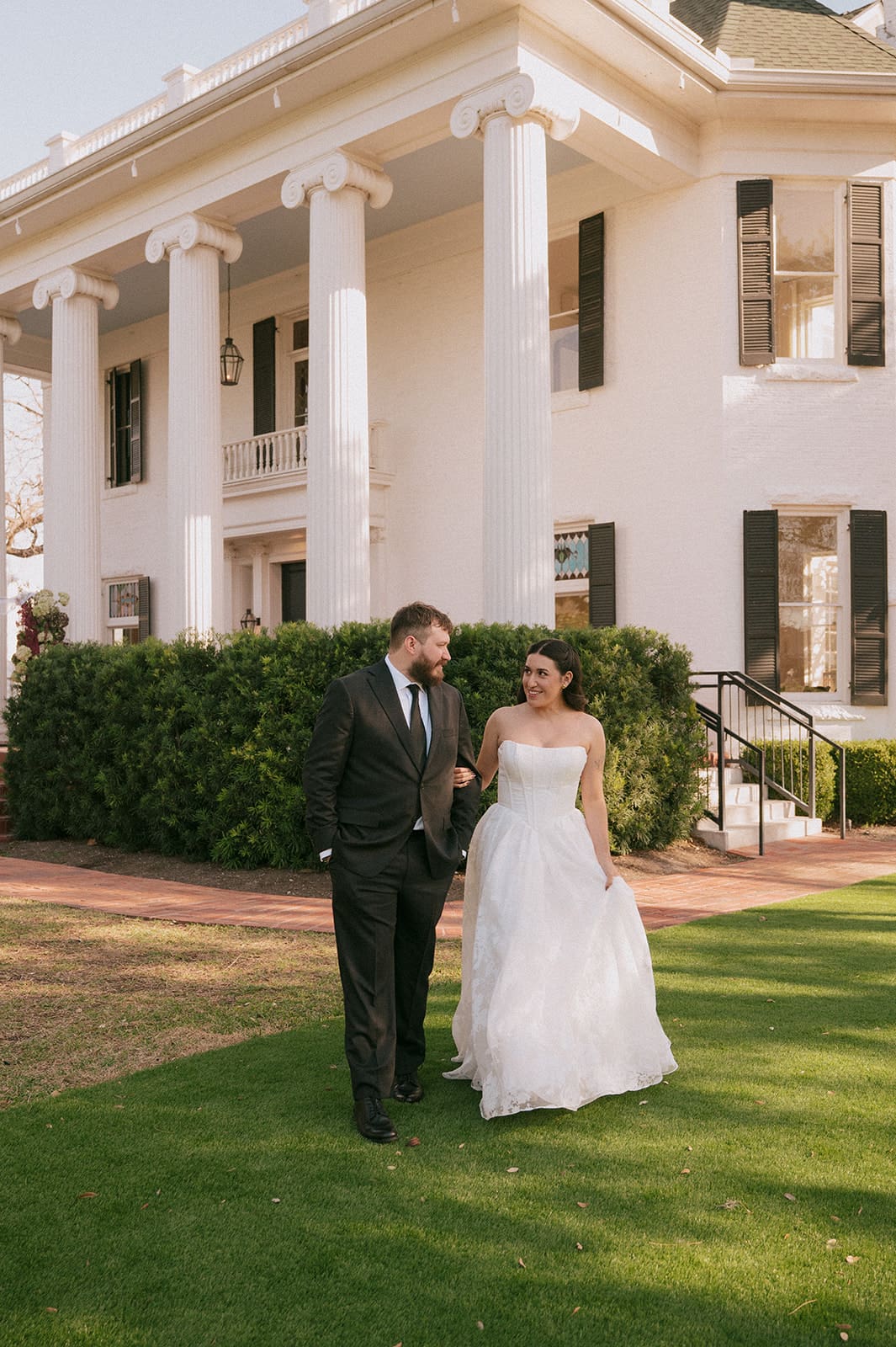 A man in a black suit and a woman in a white wedding dress stand side by side outdoors in front of the woodbine mansion
