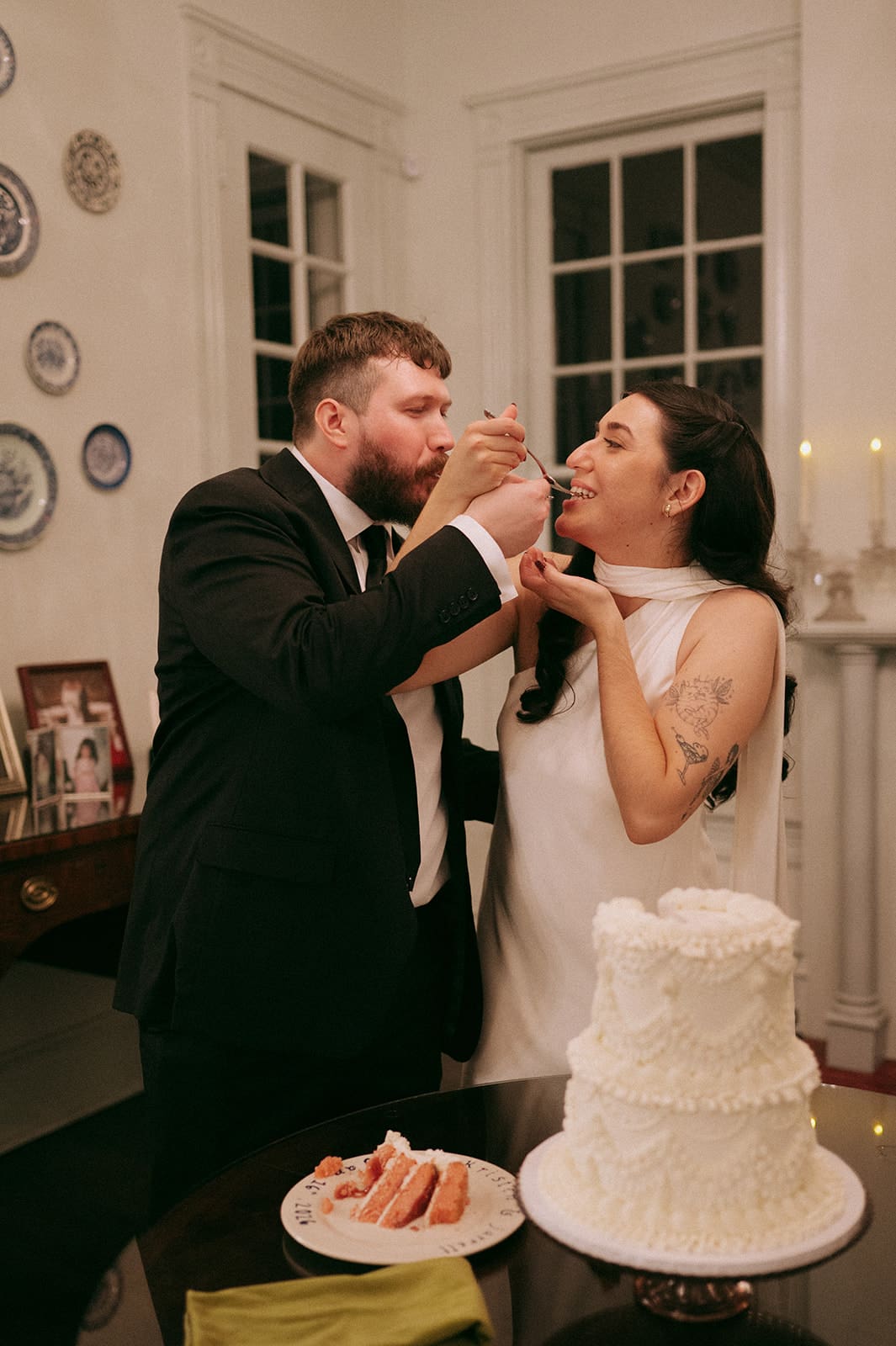 A couple in formal attire stands together cutting a white, three-tiered cake on a table in a room with white walls and large windows at Woodbine Mansion