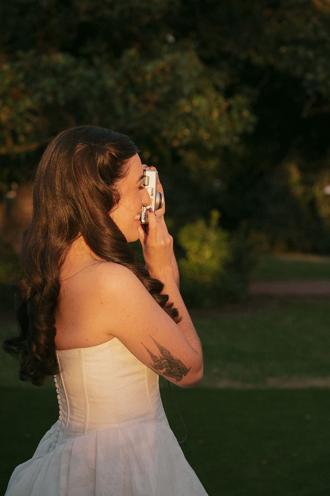 A woman in a strapless white dress stands outdoors, smiling and holding a camera to her face, with trees and grass in the background.