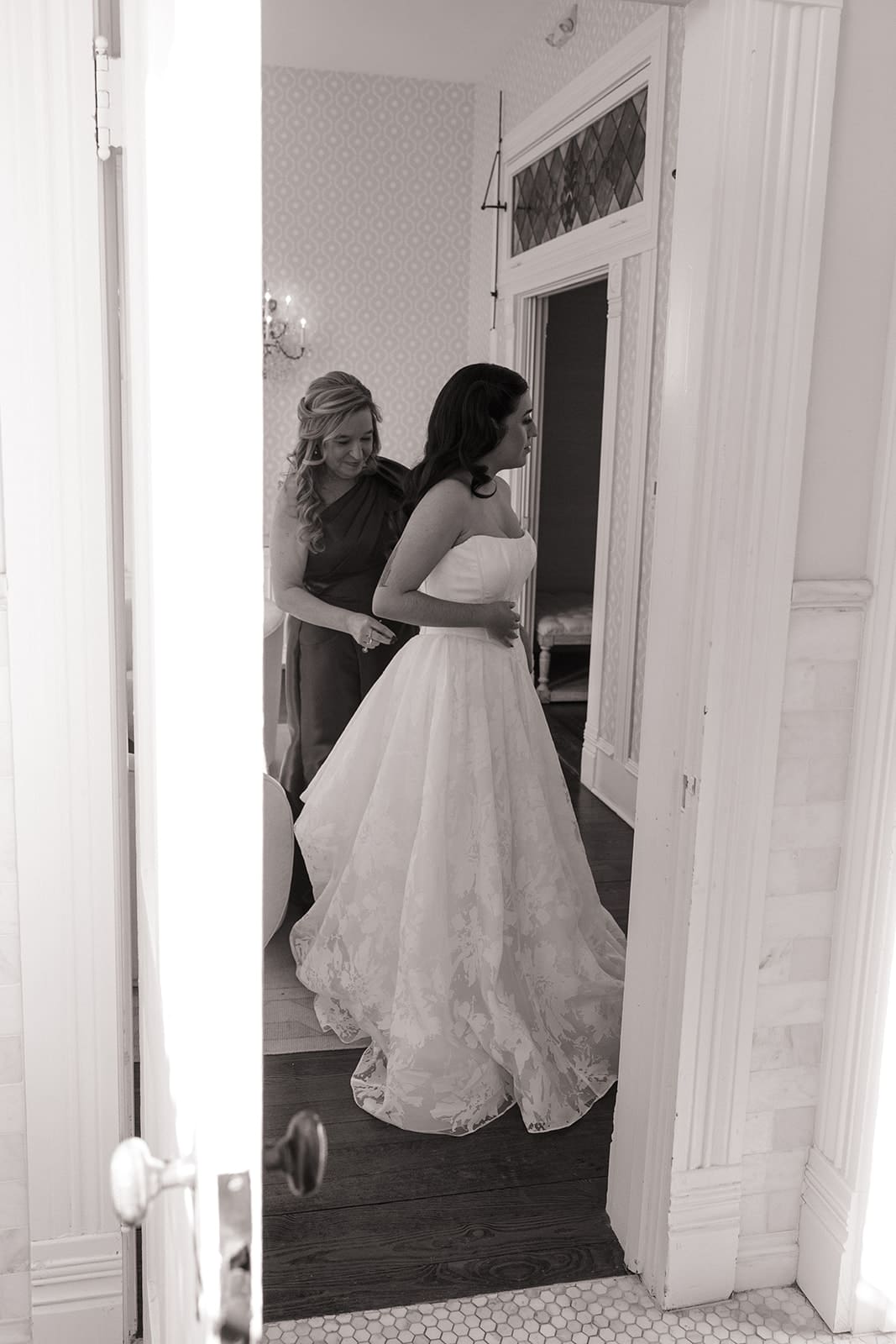 A woman in a wedding dress stands as another woman helps adjust the back of her dress in a well-lit room, seen through a partially open door at Woodbine Mansion
