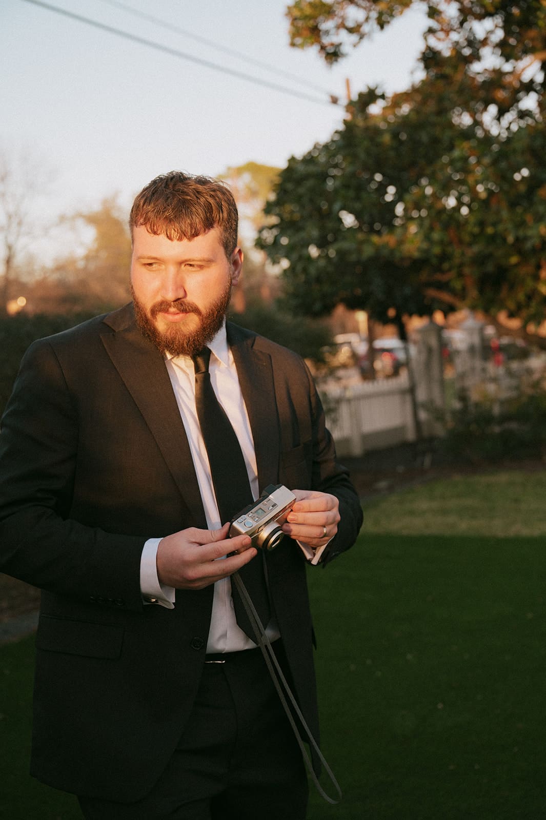 A man in a dark suit and tie stands outdoors on grass, holding a camera in his hands with trees and a fence in the background.