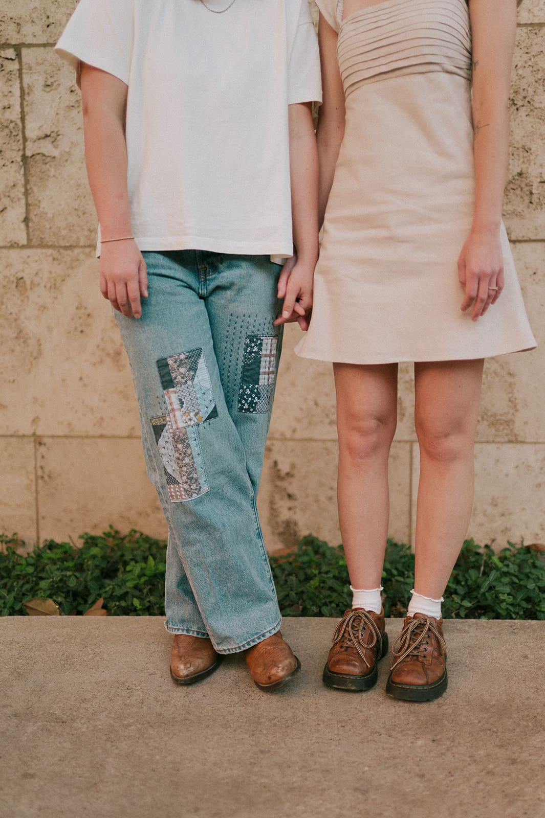 Two people walk side by side on a sidewalk next to a stone wall, smiling at each other. One wears a light dress, the other wears a white shirt and jeans.