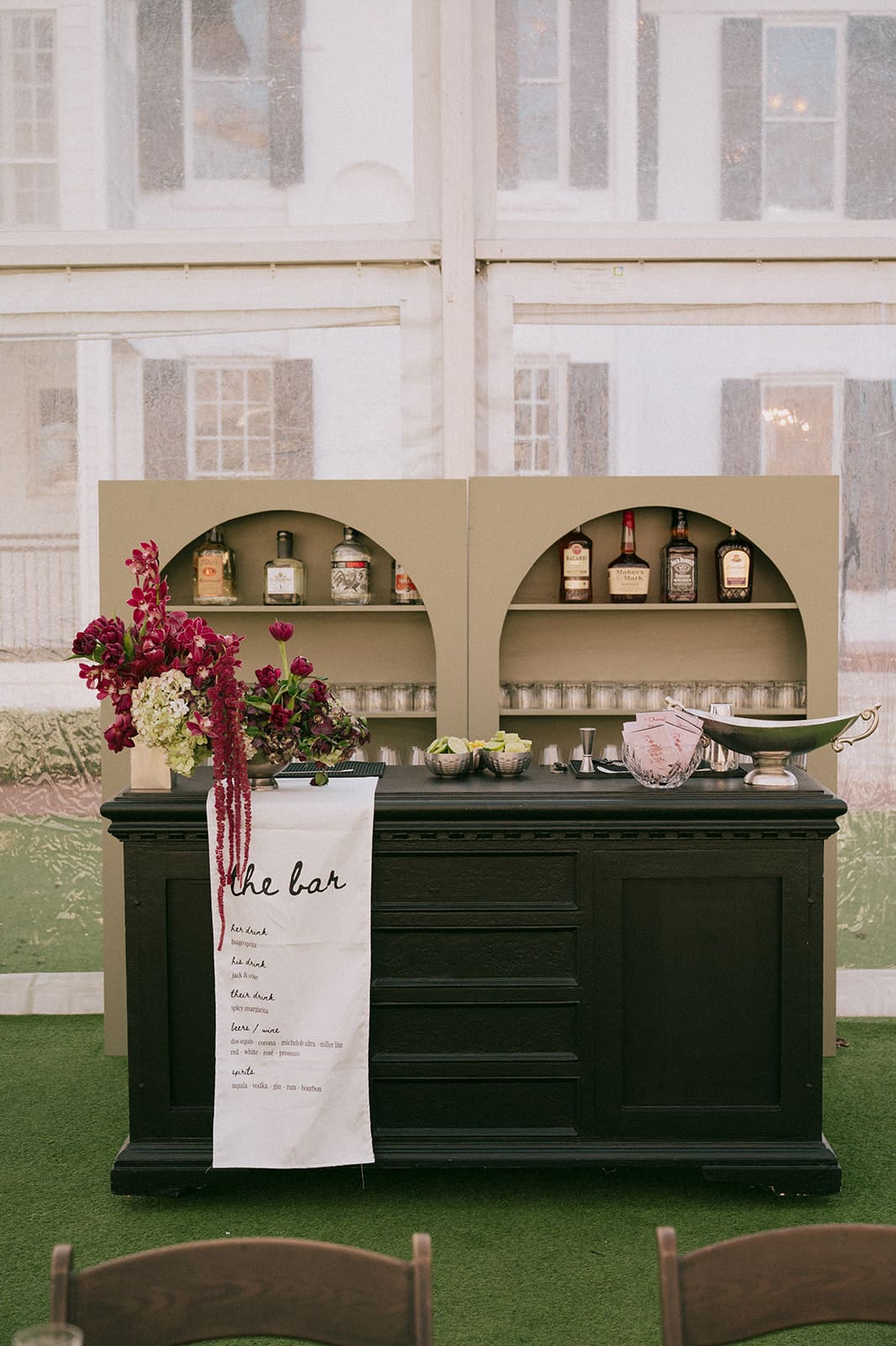 A black bar with a flower arrangement, a drink menu, and bottles of liquor displayed on shelves behind it, set indoors with large windows in the background at Woodbine Mansion