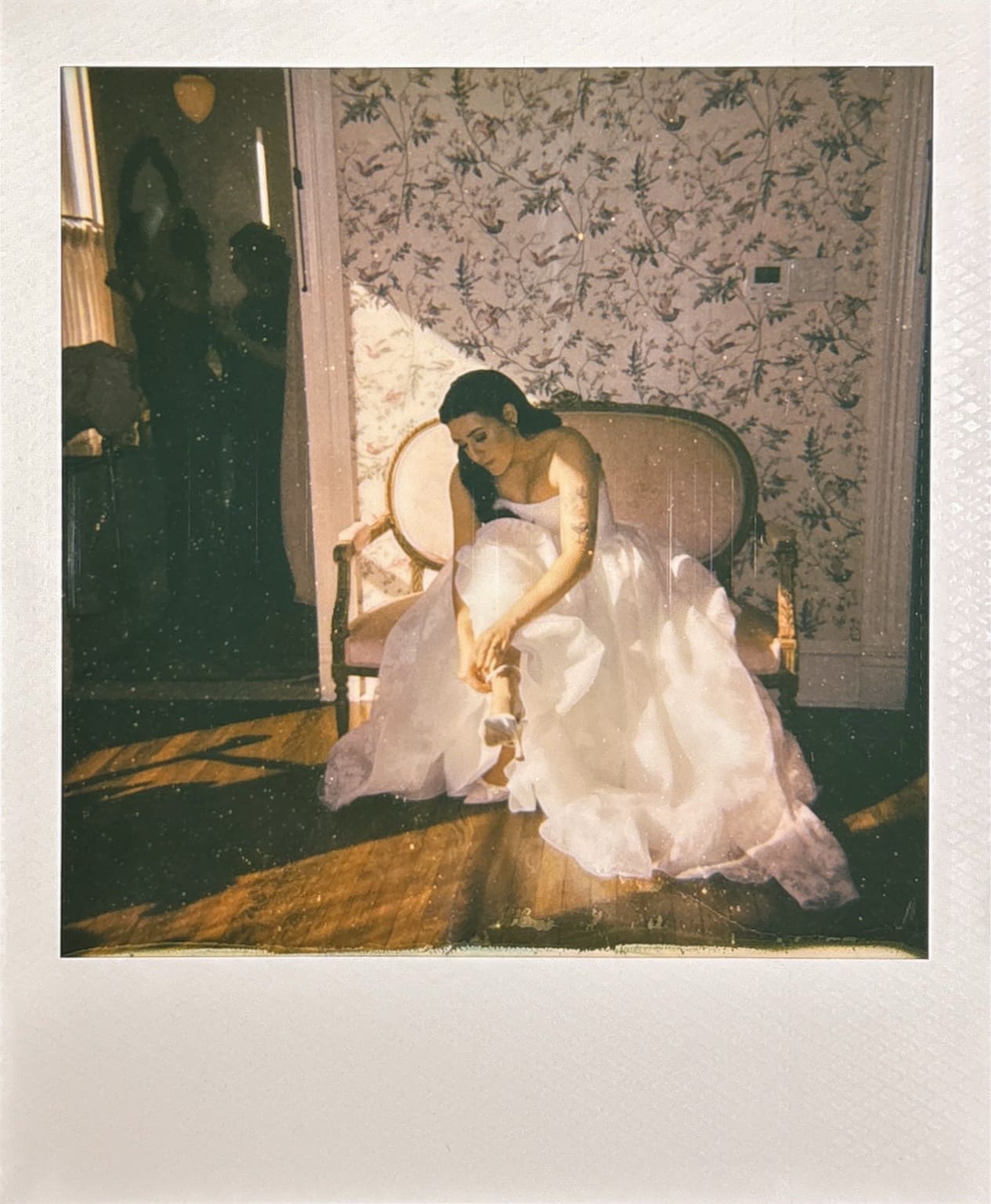 A woman in a white dress sits on a sofa in a vintage room, adjusting her shoe. Sunlight streams through a window, casting shadows across the floor and wall.