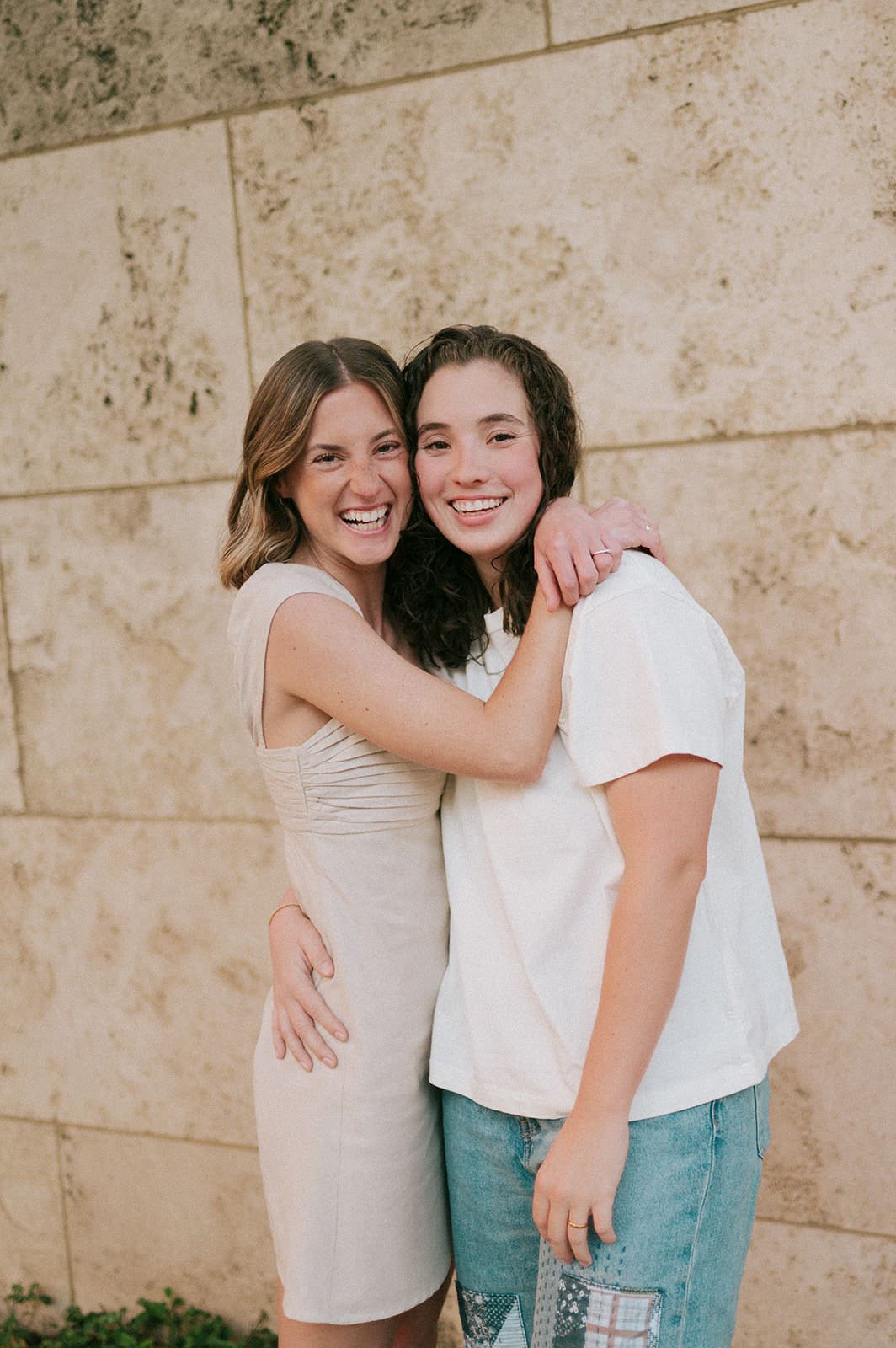 Two people walk side by side on a sidewalk next to a stone wall, smiling at each other. One wears a light dress, the other wears a white shirt and jeans.