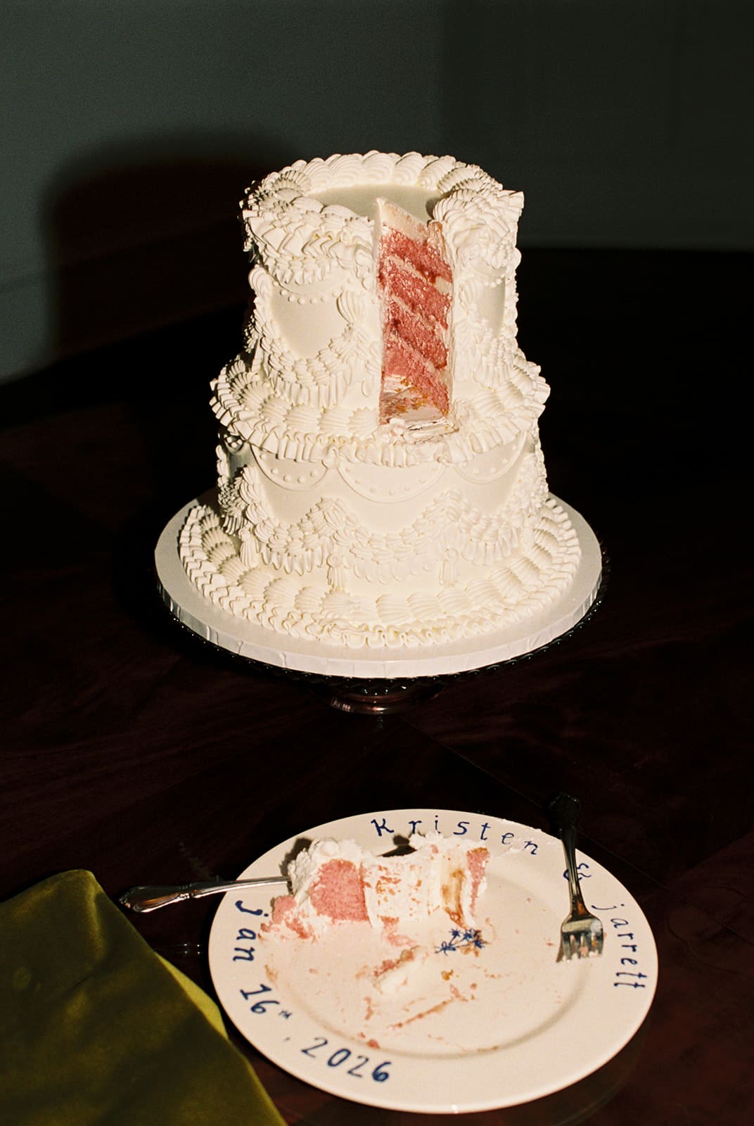 A partially eaten, two-tiered white cake with intricate frosting sits on a stand behind a plate with cake remnants and two forks.