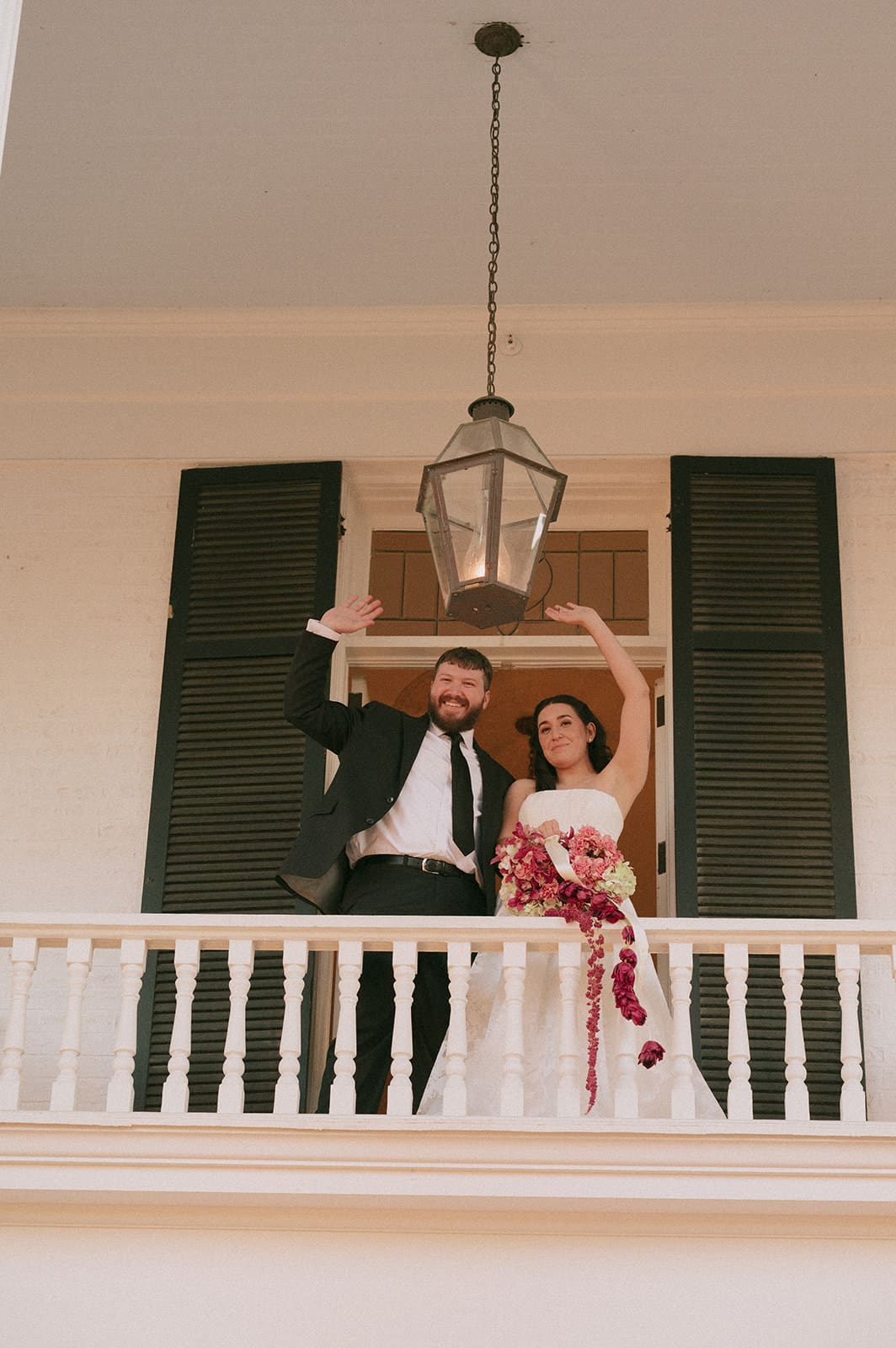A bride and groom stand on a balcony, smiling and waving. The bride holds a bouquet of red flowers, and both are dressed in formal wedding attire.
