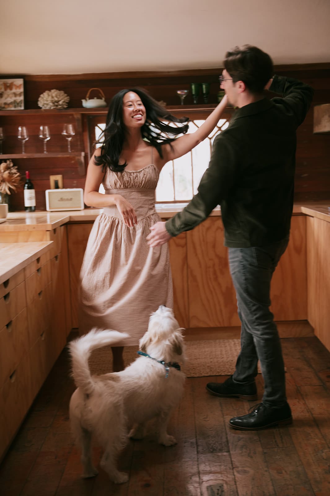 A woman and man dance together in a kitchen while a white dog stands nearby, looking up at them for their unique engagement photos