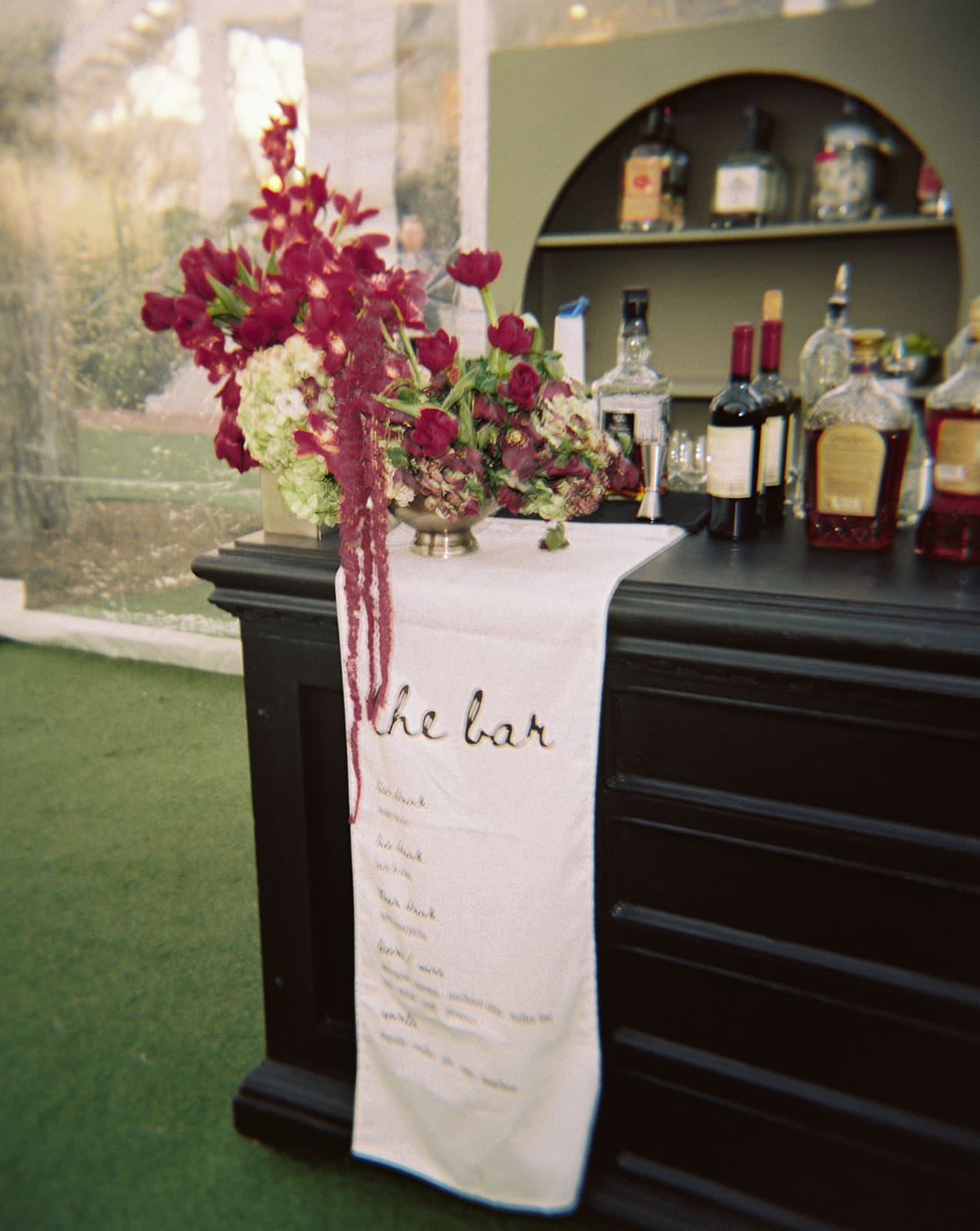 A black bar with assorted liquor bottles, a vase of red flowers, and a white cloth menu labeled “the bar” draped over the counter at Woodbine Mansion