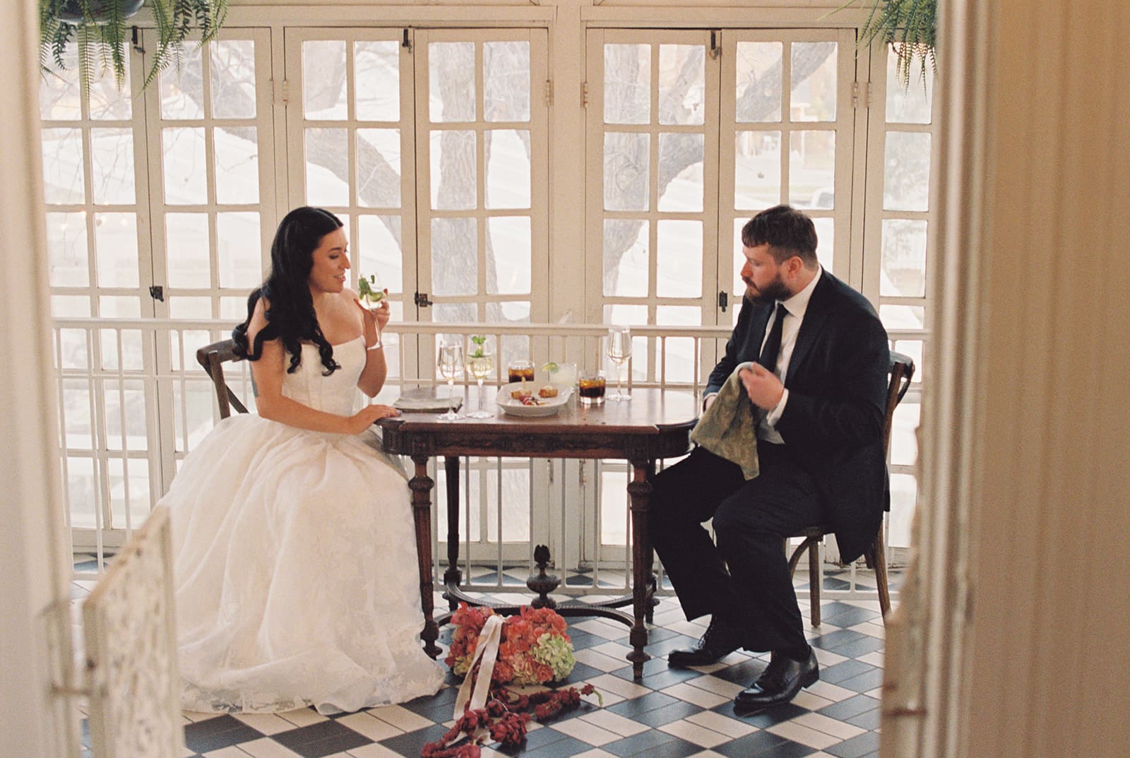 A bride and groom sit across from each other at a small table in a sunlit room, with bridal flowers on the floor and food on the table.