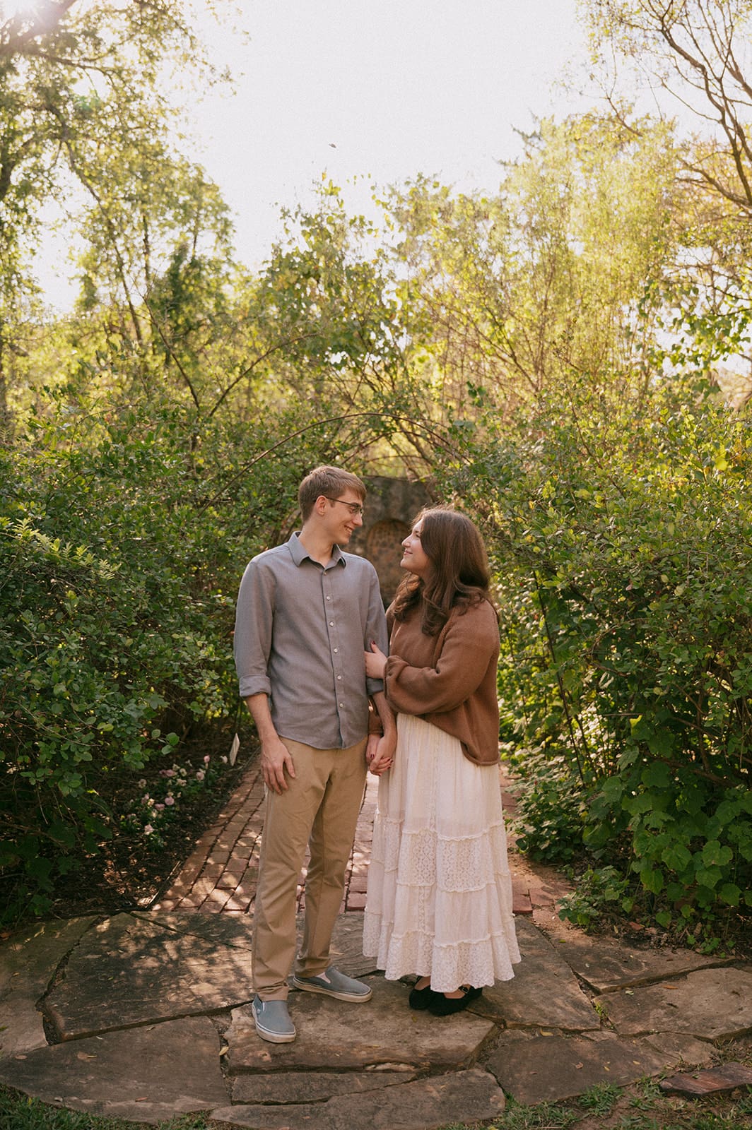 A couple stands together on a stone path in a garden, with trees and greenery in the background. The woman is smiling while the man stands behind her, holding her waist.