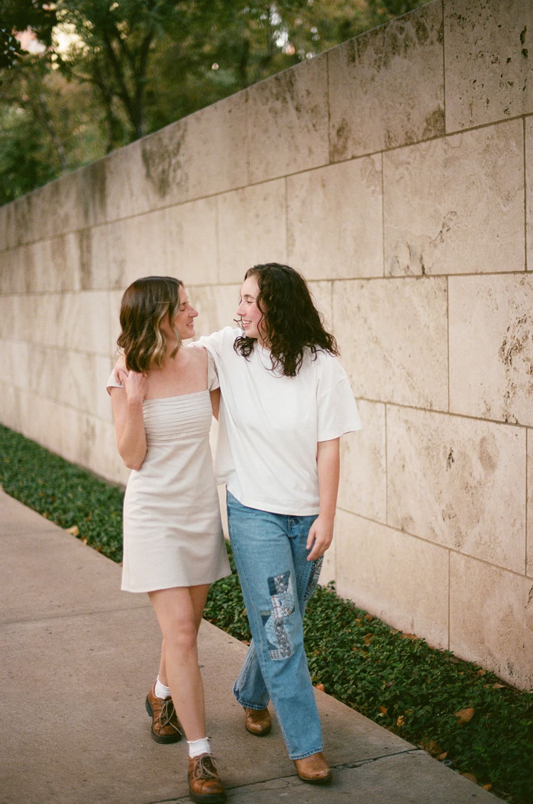 Two people walk side by side on a sidewalk next to a stone wall, smiling at each other. One wears a light dress, the other wears a white shirt and jeans.