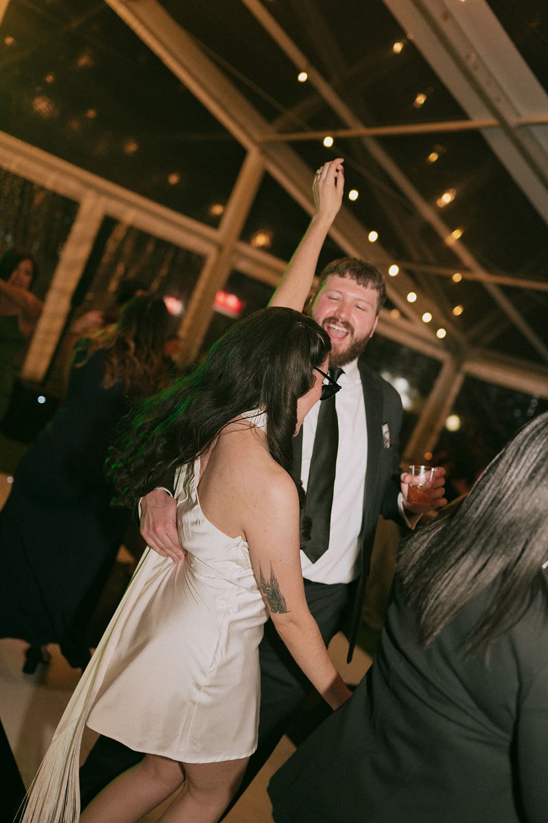 A man in a suit and a woman in a white dress dance together at an indoor event. The man raises one arm while holding a drink in the other. Other people are visible in the background.