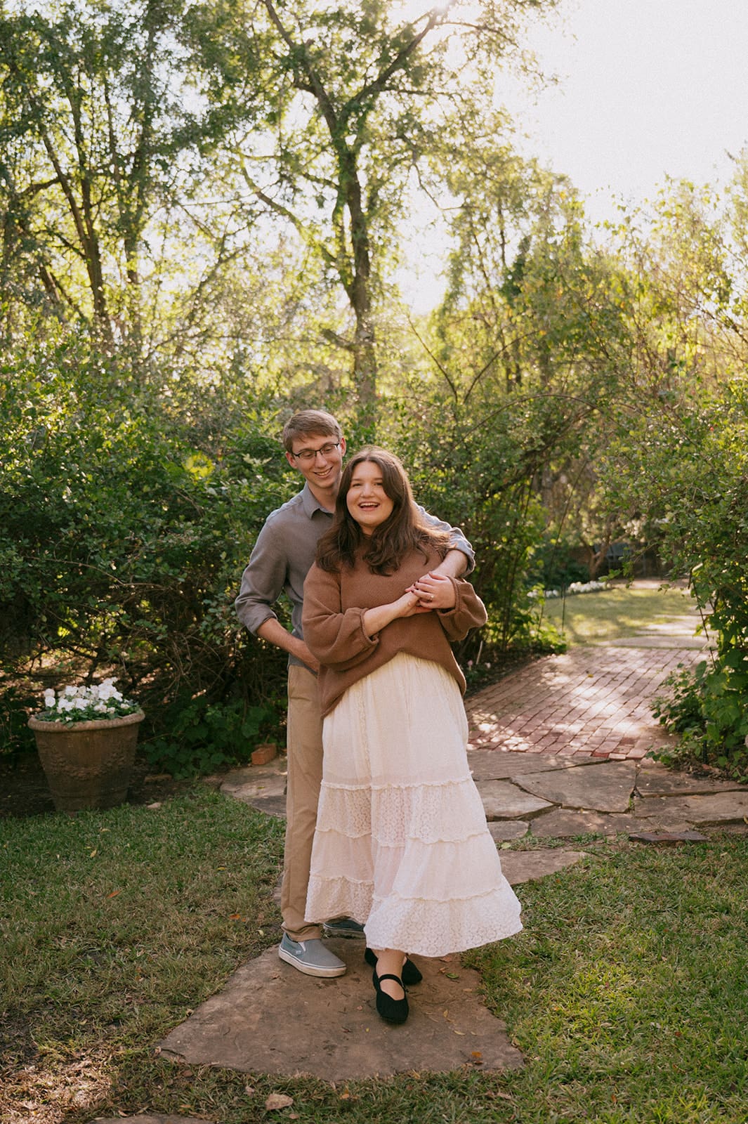 A couple stands together on a stone path in a garden, with trees and greenery in the background. The woman is smiling while the man stands behind her, holding her waist.