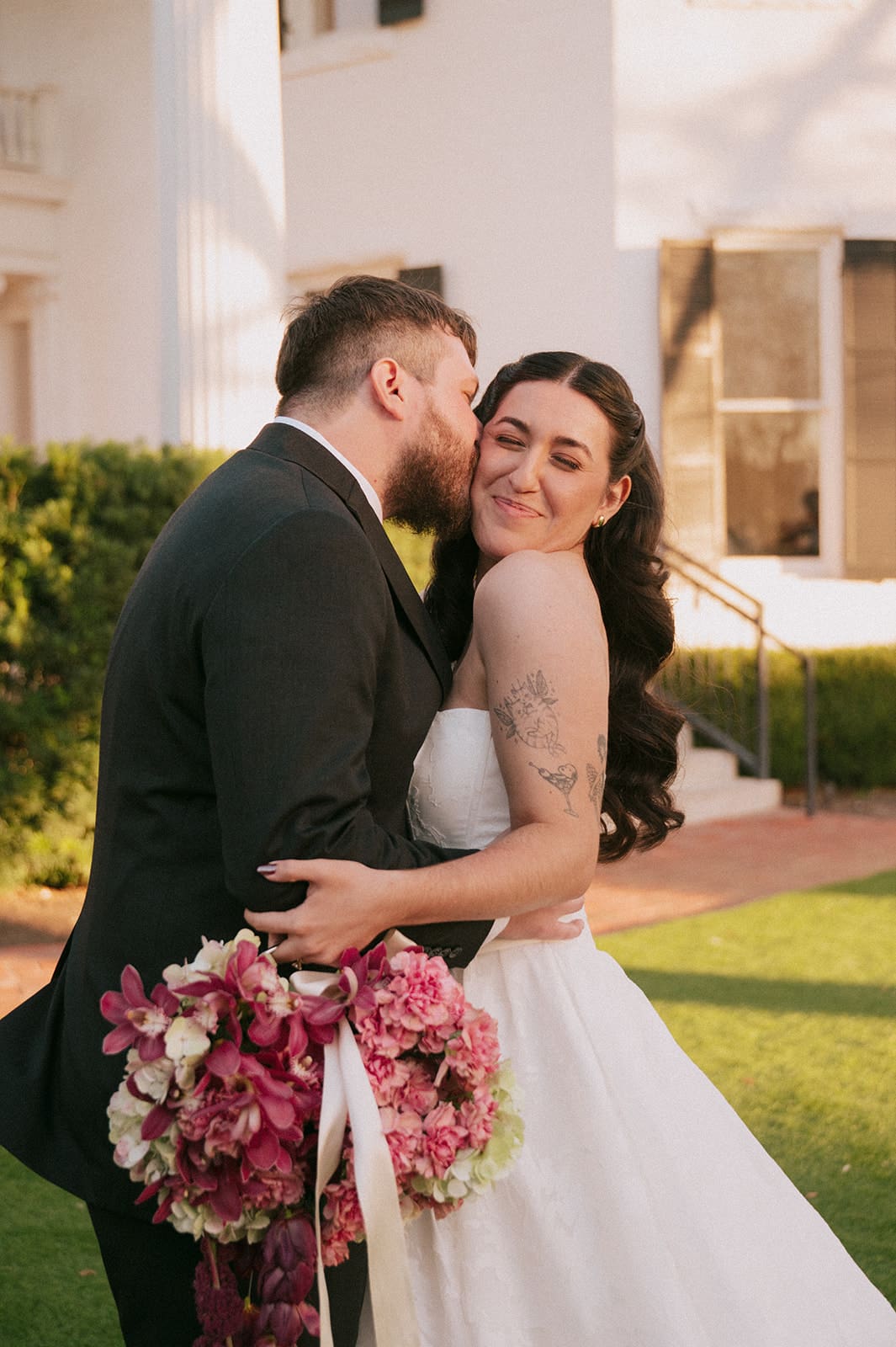 A groom in a black suit kisses a smiling bride in a white dress holding a bouquet of pink flowers outdoors in front of the woodbine mansion