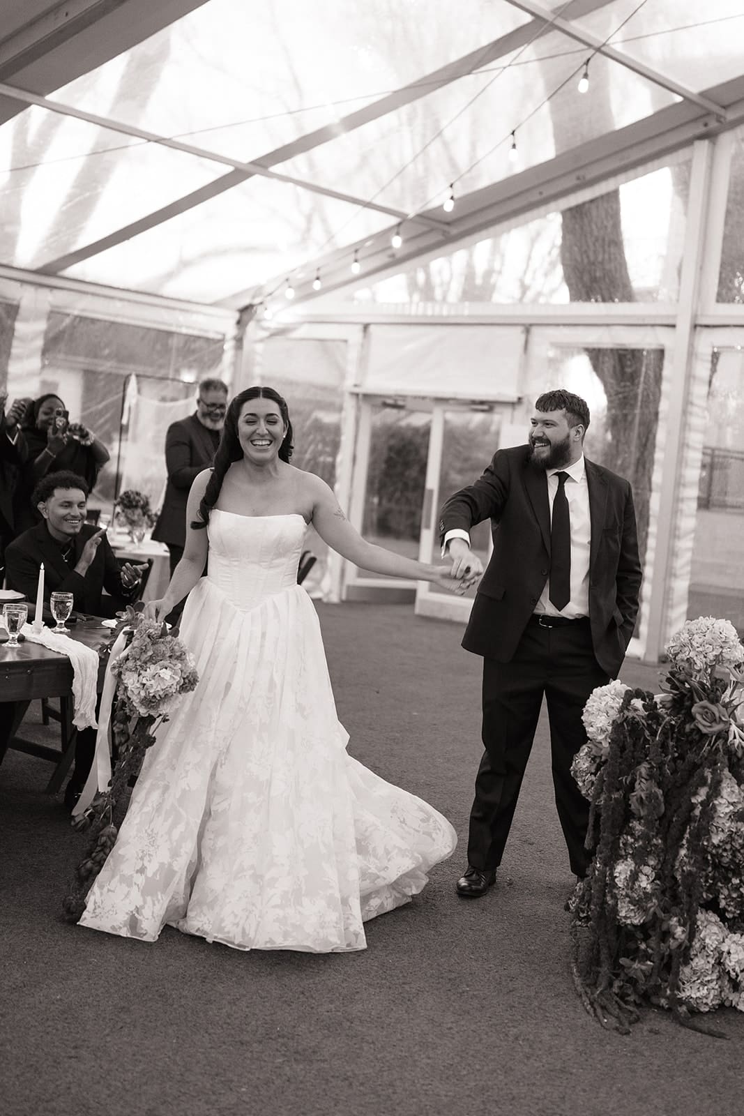 A bride and groom enter a reception area, holding hands and smiling, as guests seated at tables watch them.