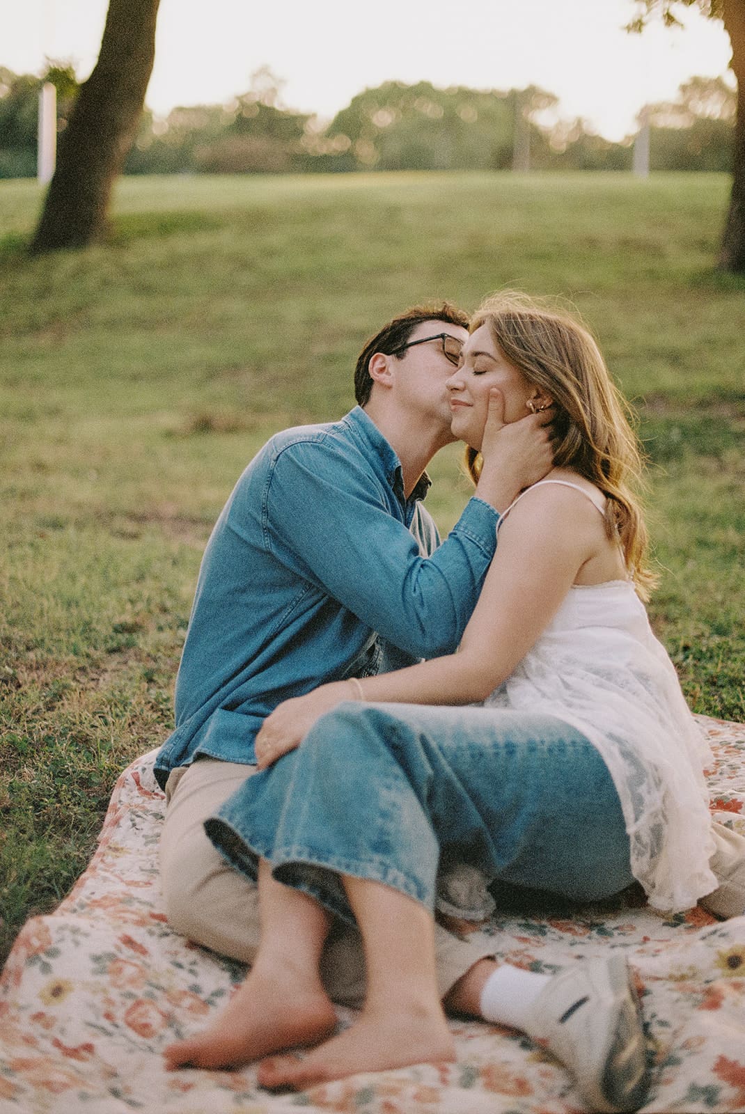 A man and a woman sit on a picnic blanket on the grass, facing each other and kissing, with a hill and trees in the background for unique engagement pictures