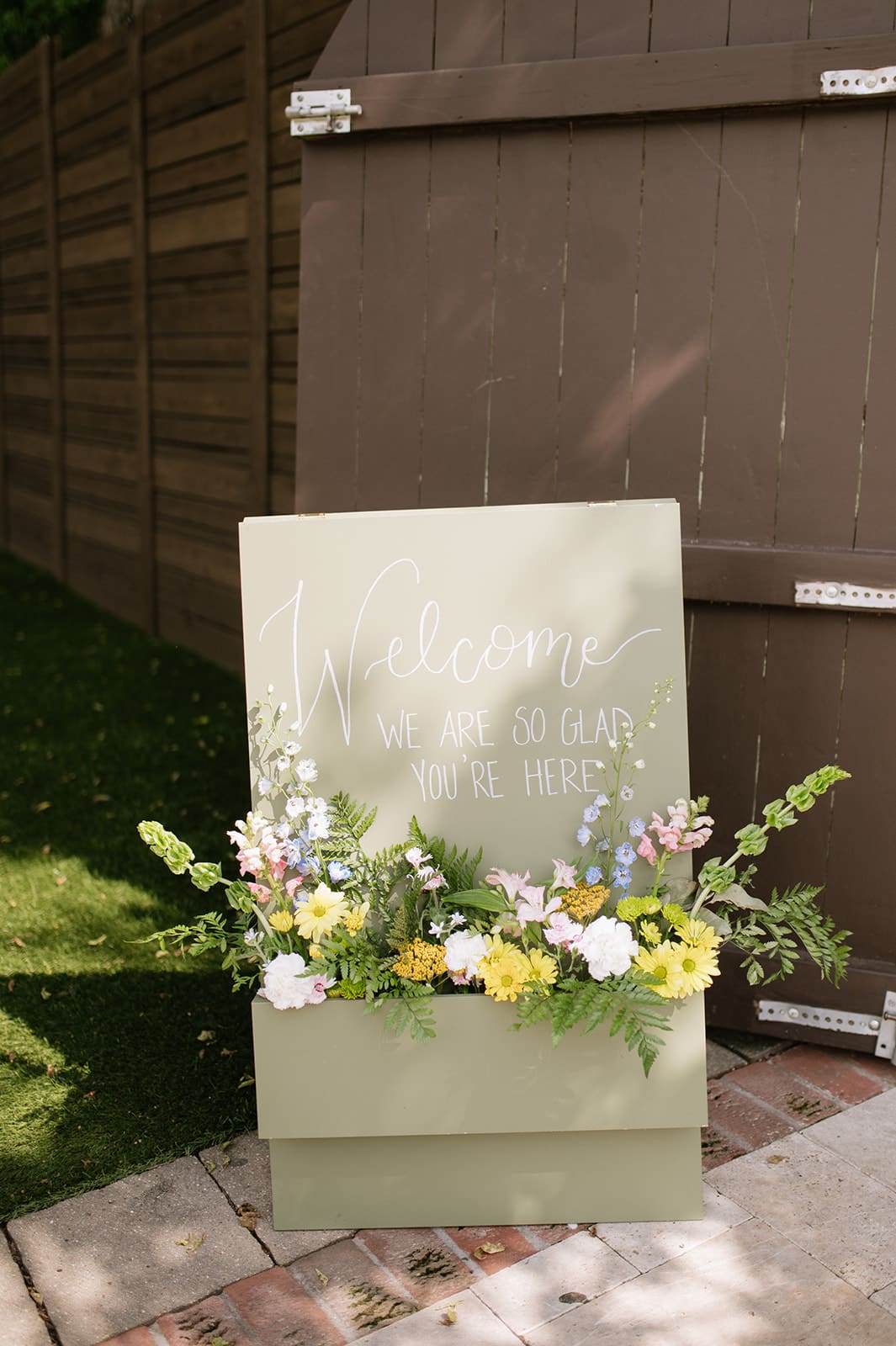 A light green welcome sign with white cursive text and a floral arrangement at the base stands outdoors on a paved area near a wooden fence.