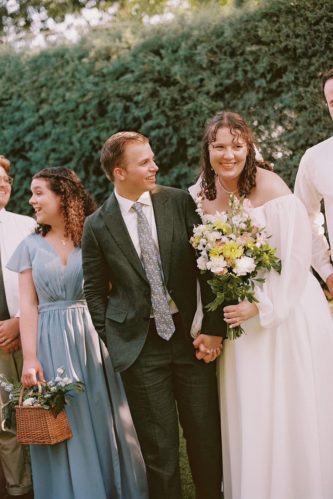 A group of six people, including a bride in a white dress and groom in a green suit, pose together outdoors, smiling at the camera at a wedding at Magnolia Terrace in Frisco, Tx