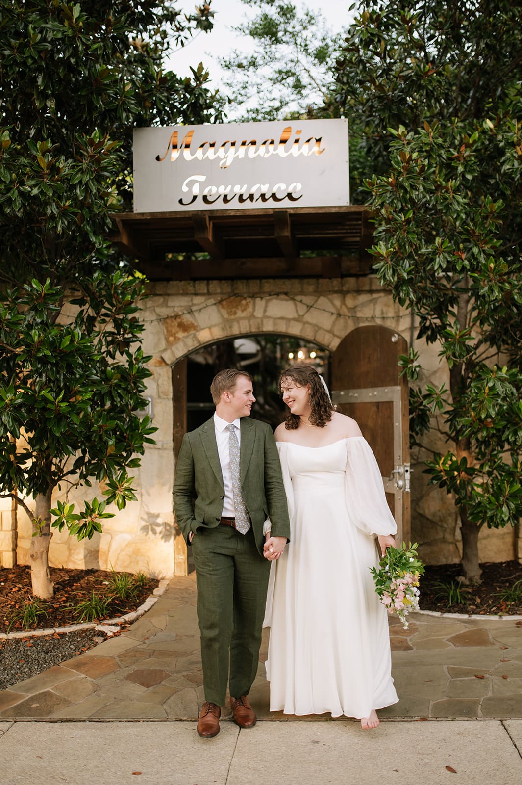 Two people in wedding attire stand close together under a veil in a garden at sunset, with light streaming through the trees.