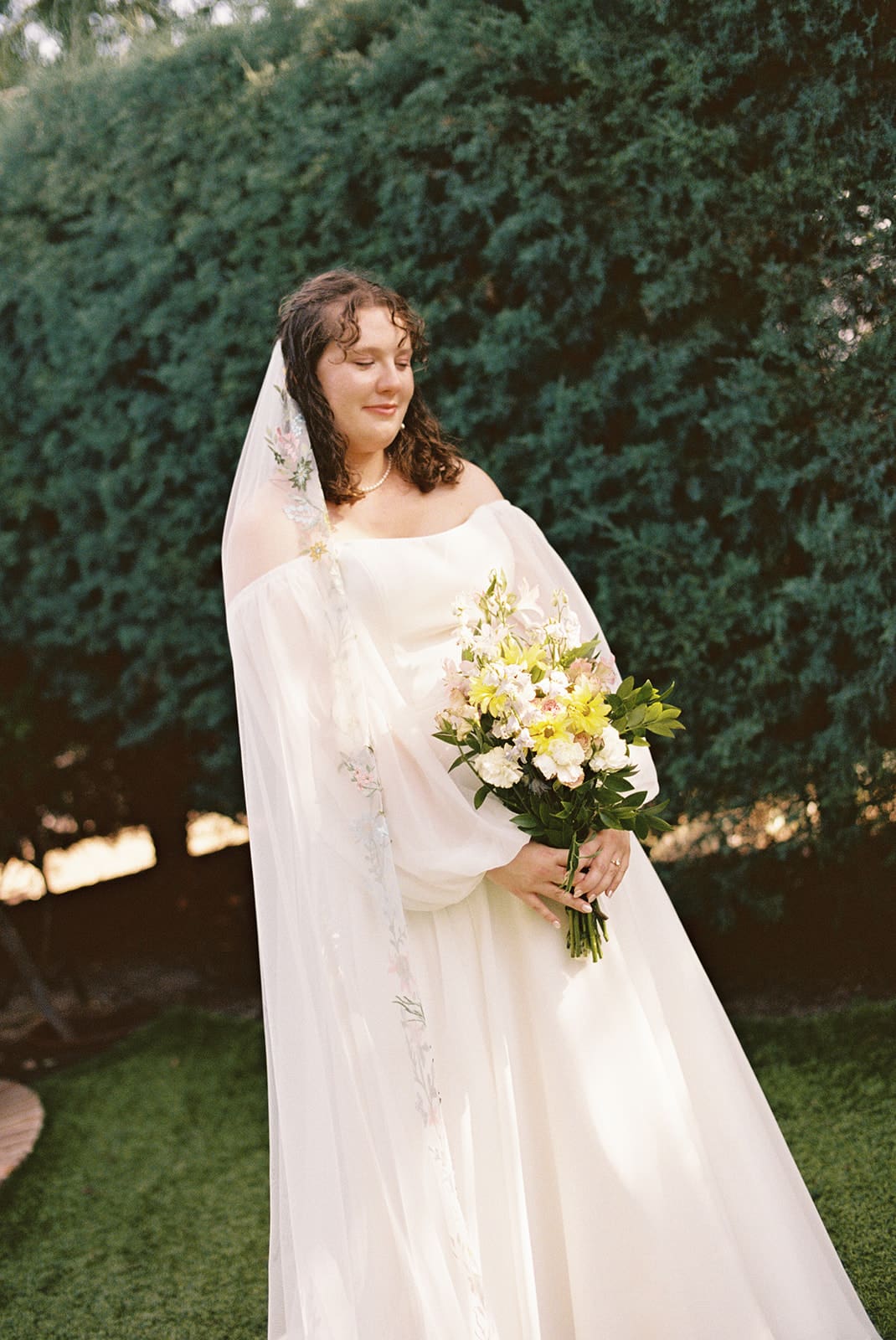 A bride in a white dress and veil stands outdoors holding a bouquet of yellow and white flowers, with greenery in the background.