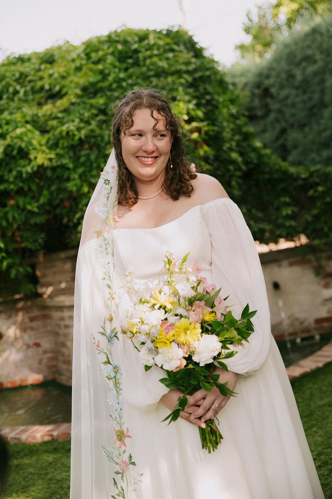 A bride in a white dress and veil stands outdoors holding a bouquet of yellow and white flowers, with greenery in the background.
