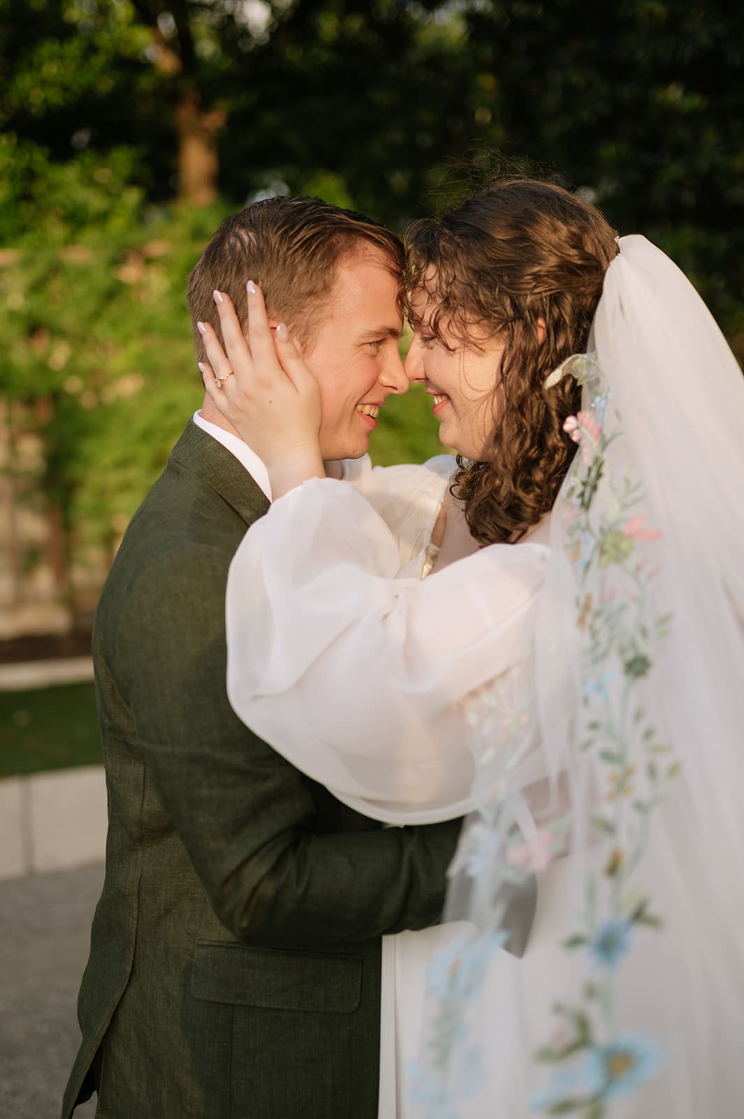 Two people in wedding attire stand close together under a veil in a garden at sunset, with light streaming through the trees.
