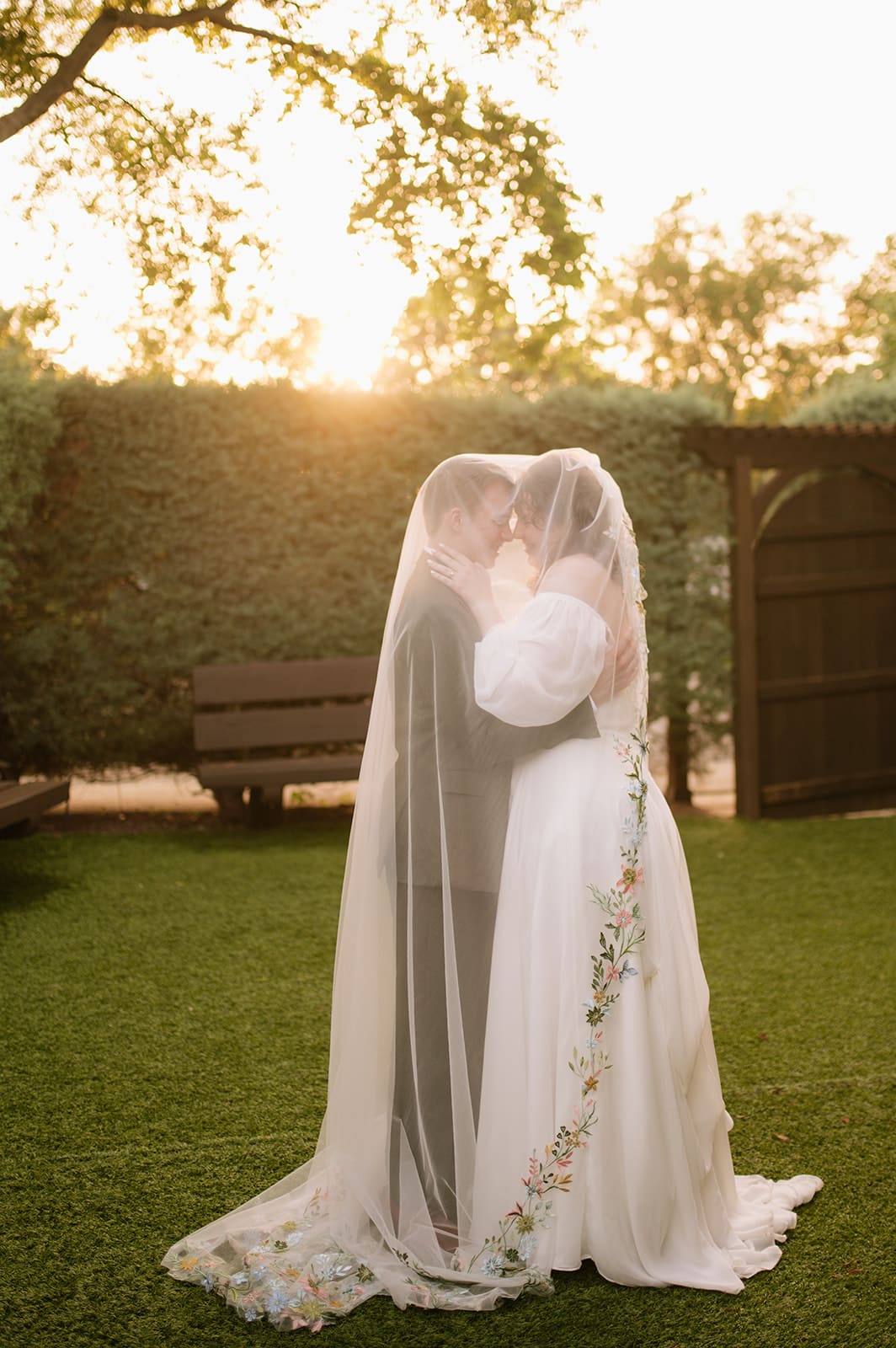 Two people in wedding attire stand close together under a veil in a garden at sunset, with light streaming through the trees.