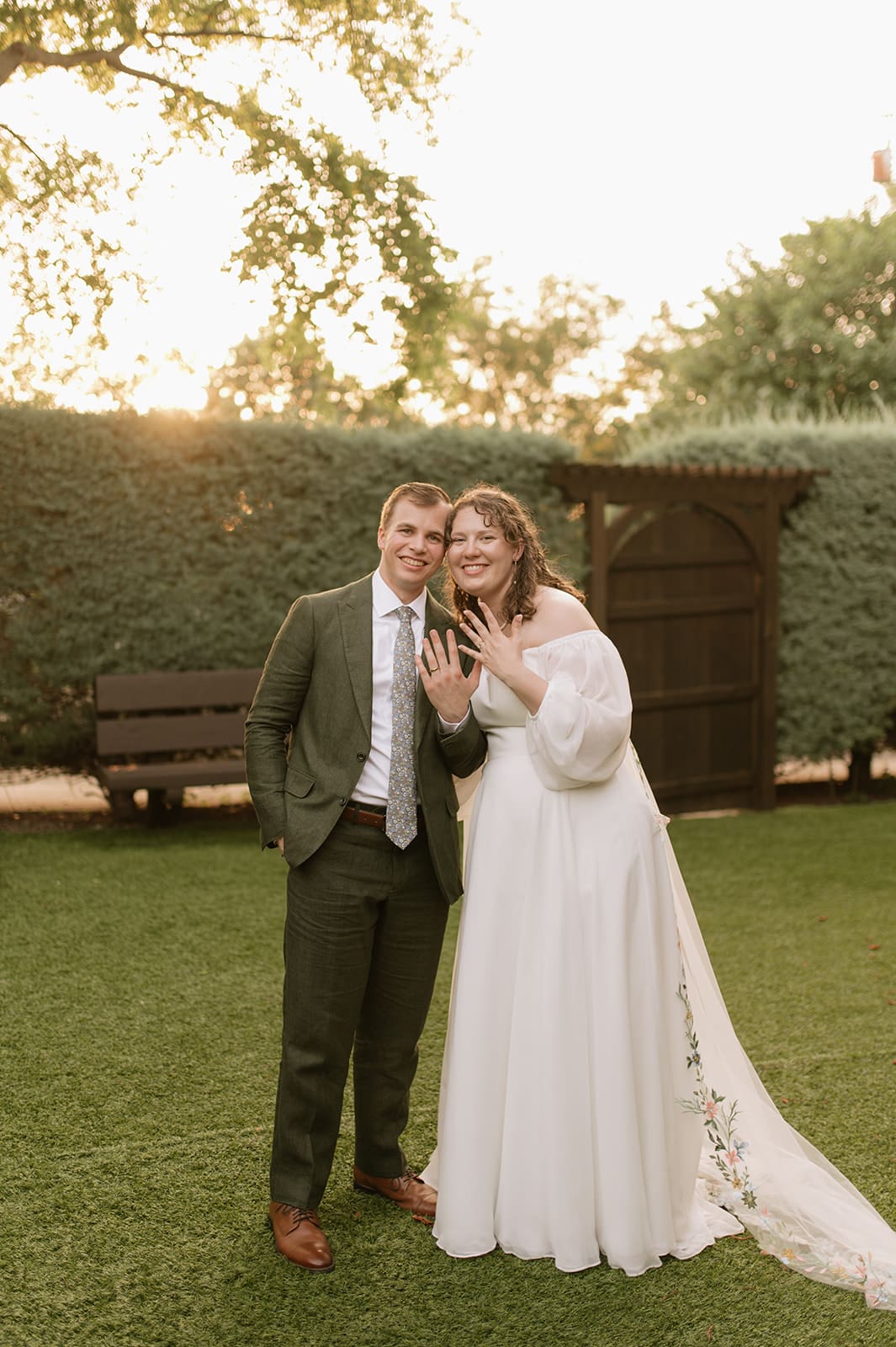 A bride in a white dress and a groom in a green suit stand together on grass, smiling and showing wedding rings, with trees and hedges in the background.