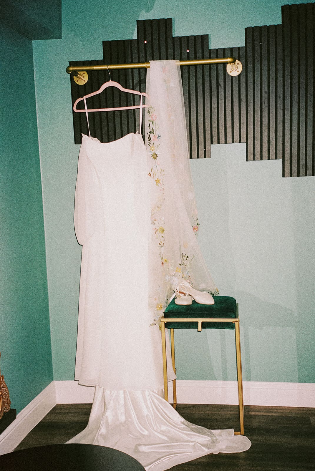 A white wedding dress on a hanger and a veil are displayed on a gold rack above a green chair, with white shoes placed on the seat.