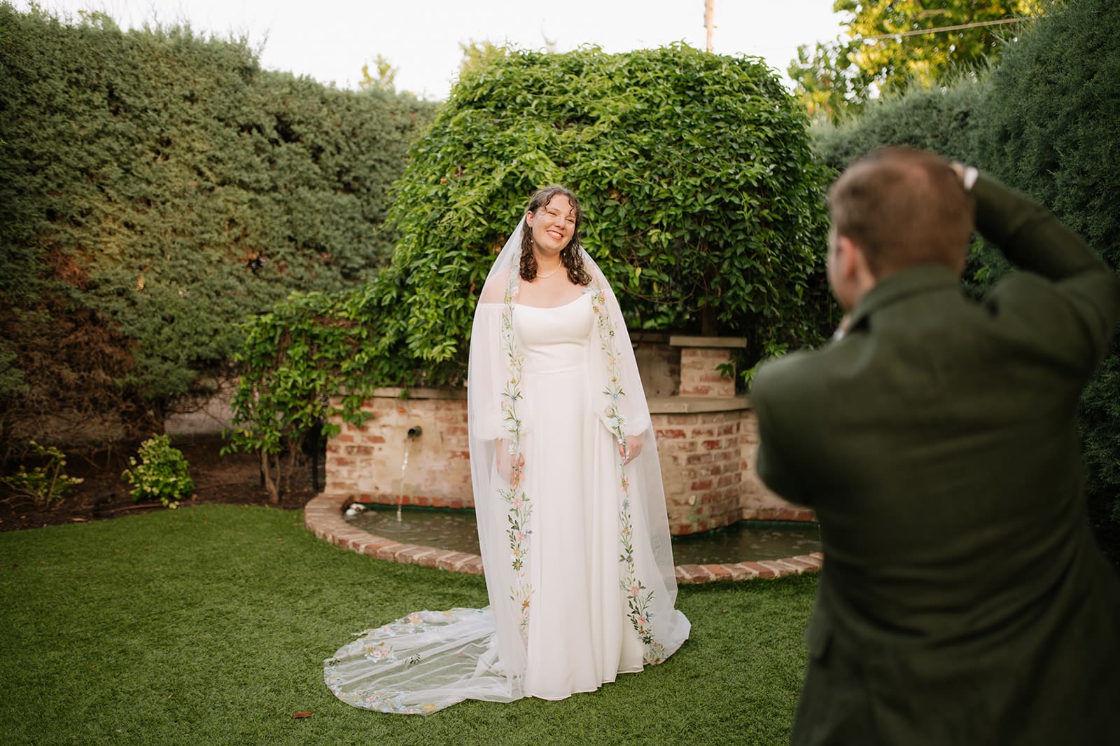 Two people in wedding attire stand close together under a veil in a garden at sunset, with light streaming through the trees.