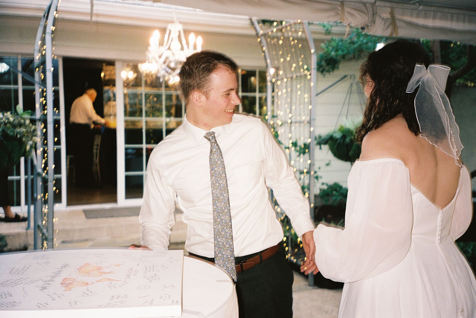 A man in a white shirt and tie holds hands with a woman in a white off-the-shoulder dress and veil at an outdoor event, with another person visible in the background.