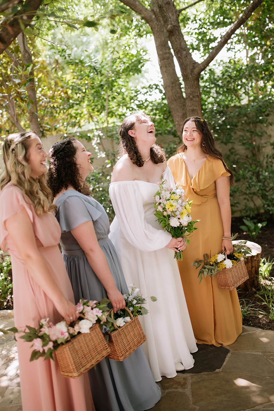Four women in colorful dresses stand outdoors, smiling and laughing. The woman in white holds a bouquet, suggesting she is the bride, while the others hold baskets of flowers.