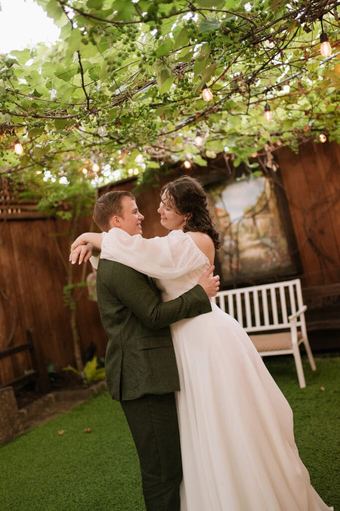 A bride and groom embrace and smile at each other under string lights in an outdoor garden setting at Magnolia Terrace in Frisco, Tx