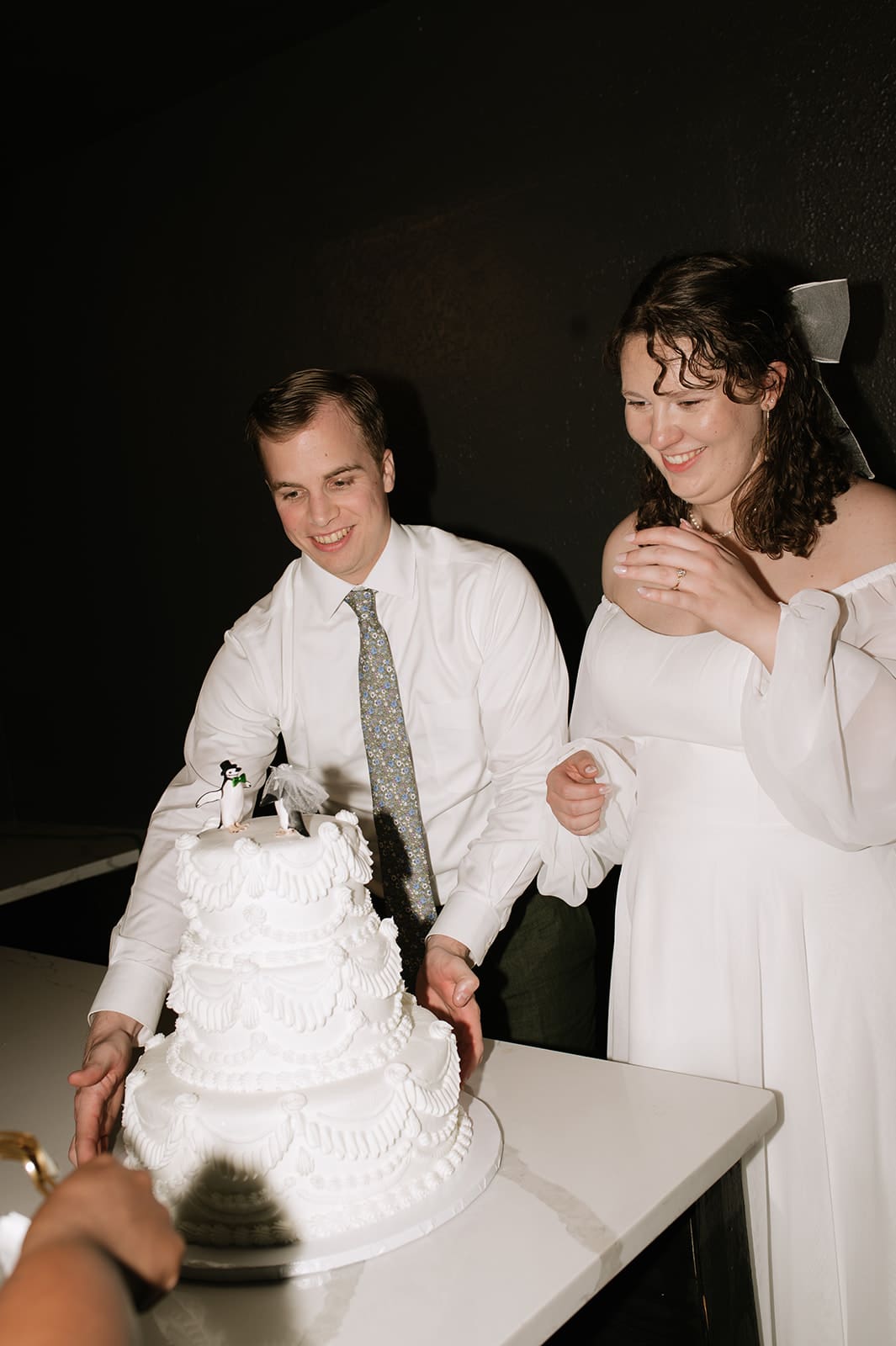 A man and woman in formal attire stand next to a white wedding cake on a table, preparing to cut it.
