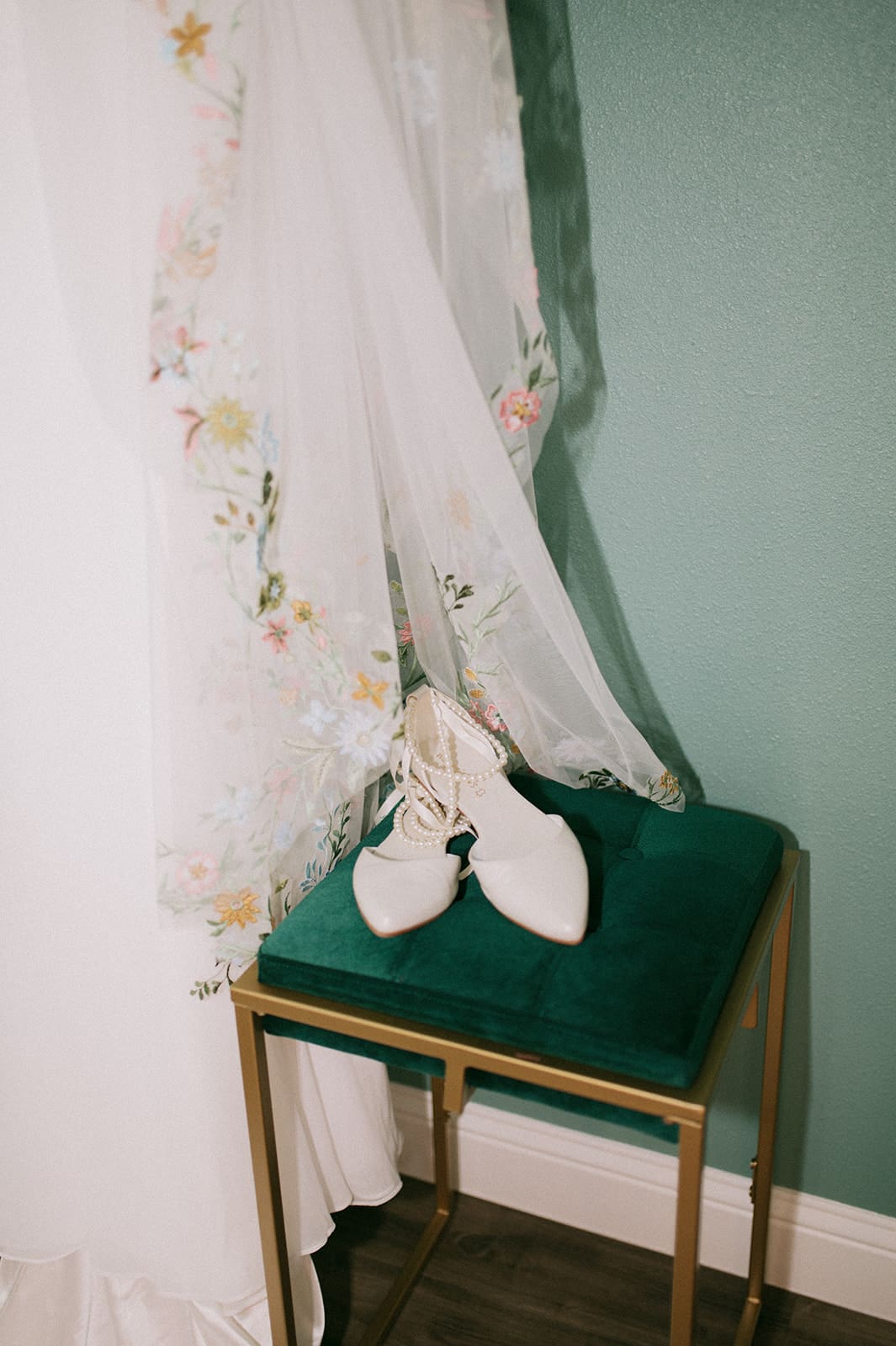 A pair of white shoes sits on a green cushioned stool next to a white curtain with floral embroidery, against a teal wall.