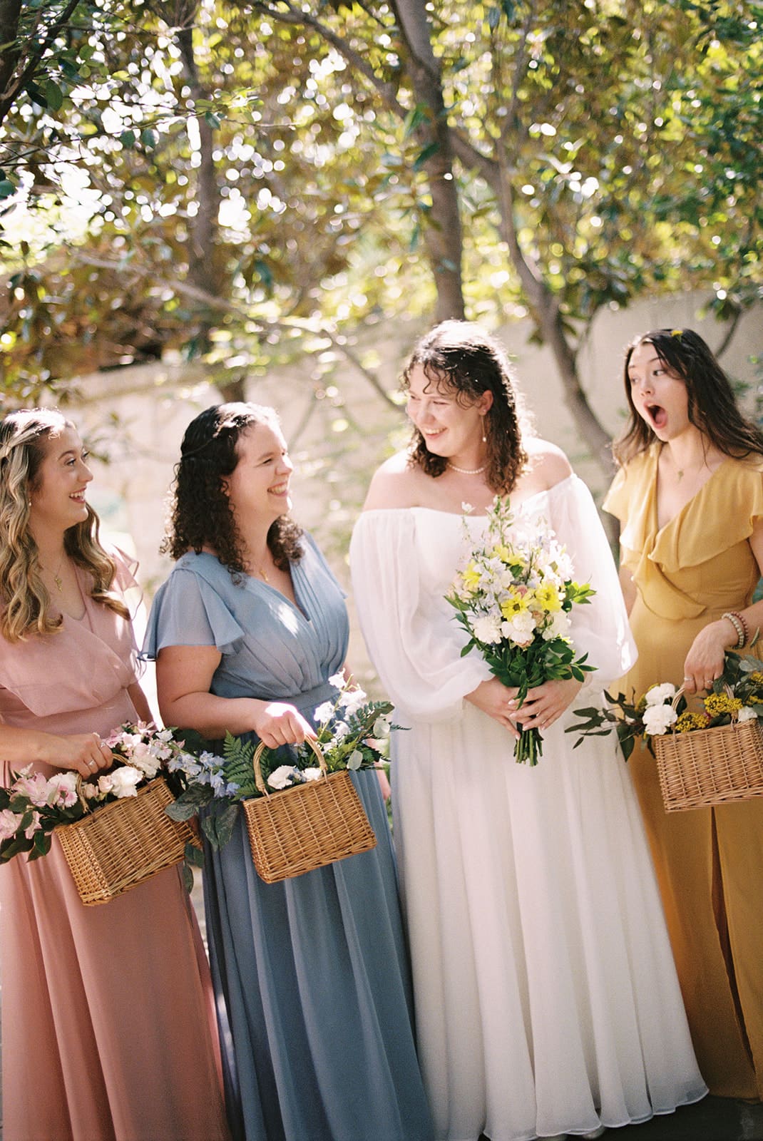 Four women in colorful dresses stand outdoors, smiling and laughing. The woman in white holds a bouquet, suggesting she is the bride, while the others hold baskets of flowers.