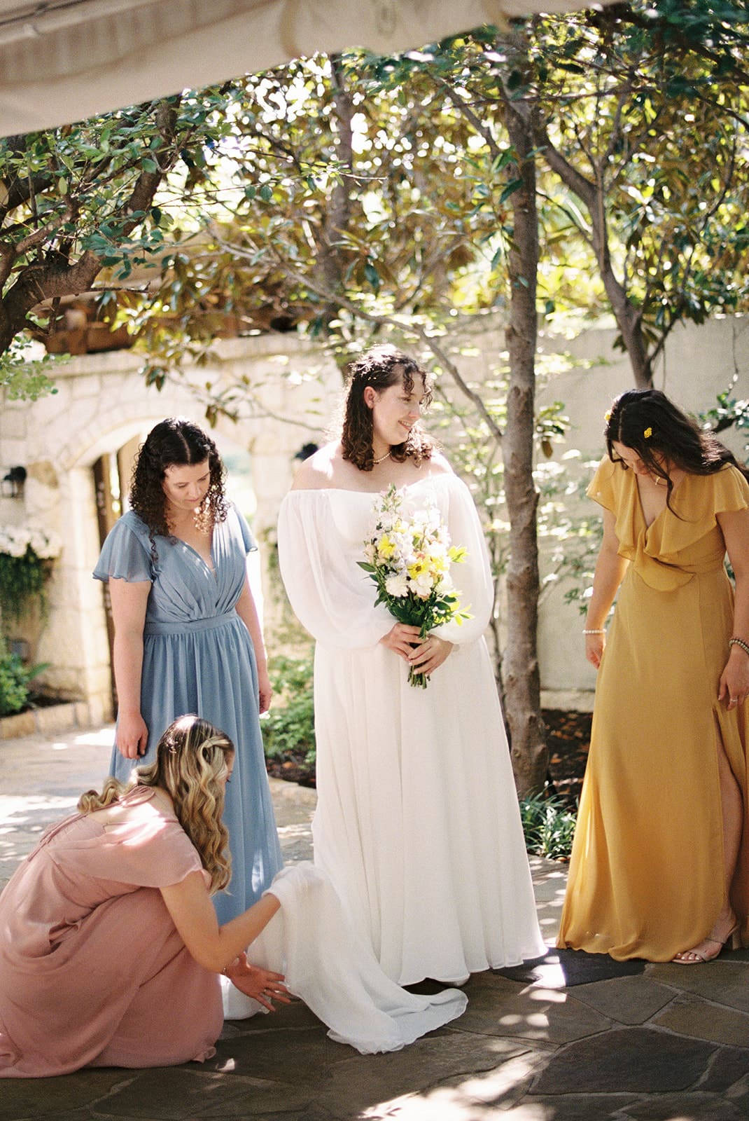 Four women in colorful dresses stand outdoors, smiling and laughing. The woman in white holds a bouquet, suggesting she is the bride, while the others hold baskets of flowers.