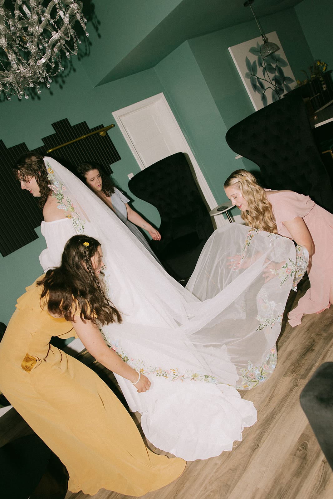 Four women in dresses adjust a bride’s wedding gown train in a room with teal walls and black chairs.