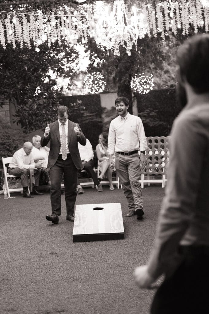 Several people gather outdoors as two men play cornhole, with string lights overhead and other guests seated in the background at Magnolia Terrace in Frisco, Tx