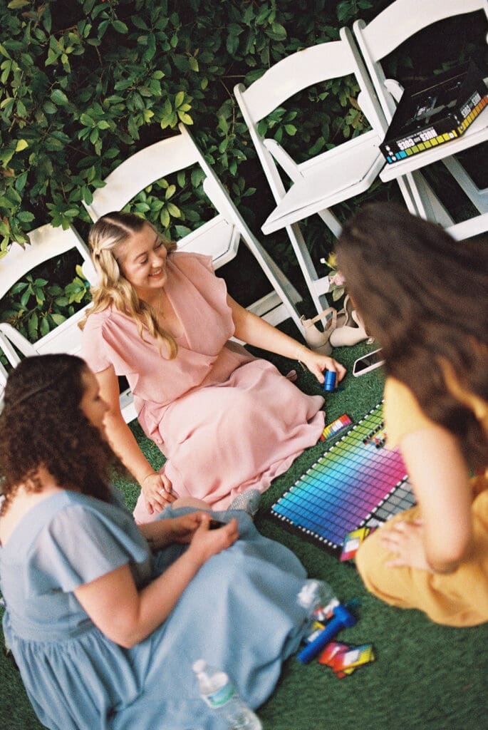 Three women sit on grass in a circle, interacting with a colorful xylophone. White folding chairs and greenery are in the background at Magnolia Terrace in Frisco, Tx