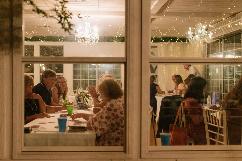 People are seated at tables inside a well-lit room, viewed through a window at night, engaged in conversation and dining.