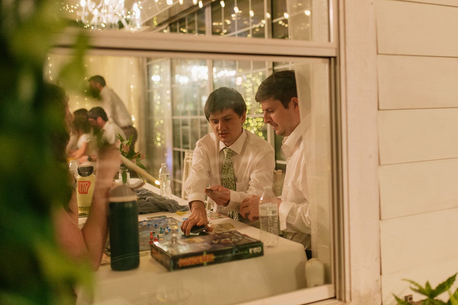 Two men in dress shirts play a board game at a table inside, seen through a window at night with string lights in the background.