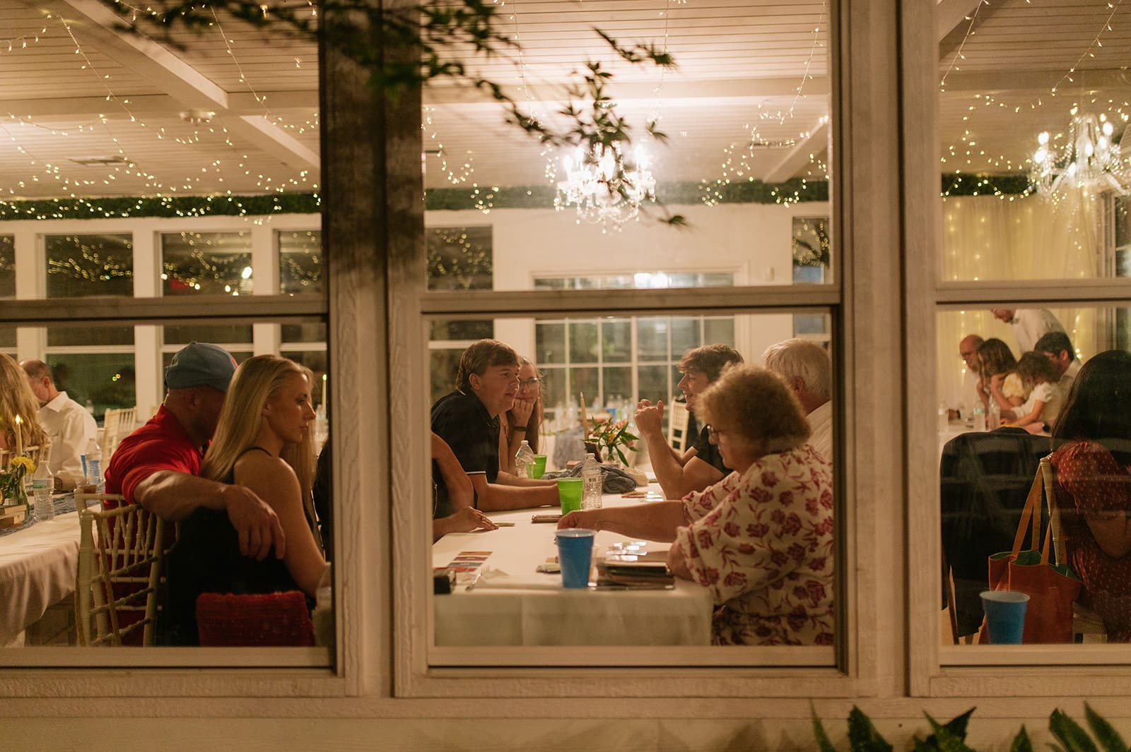 Two men in dress shirts play a board game at a table inside, seen through a window at night with string lights in the background.