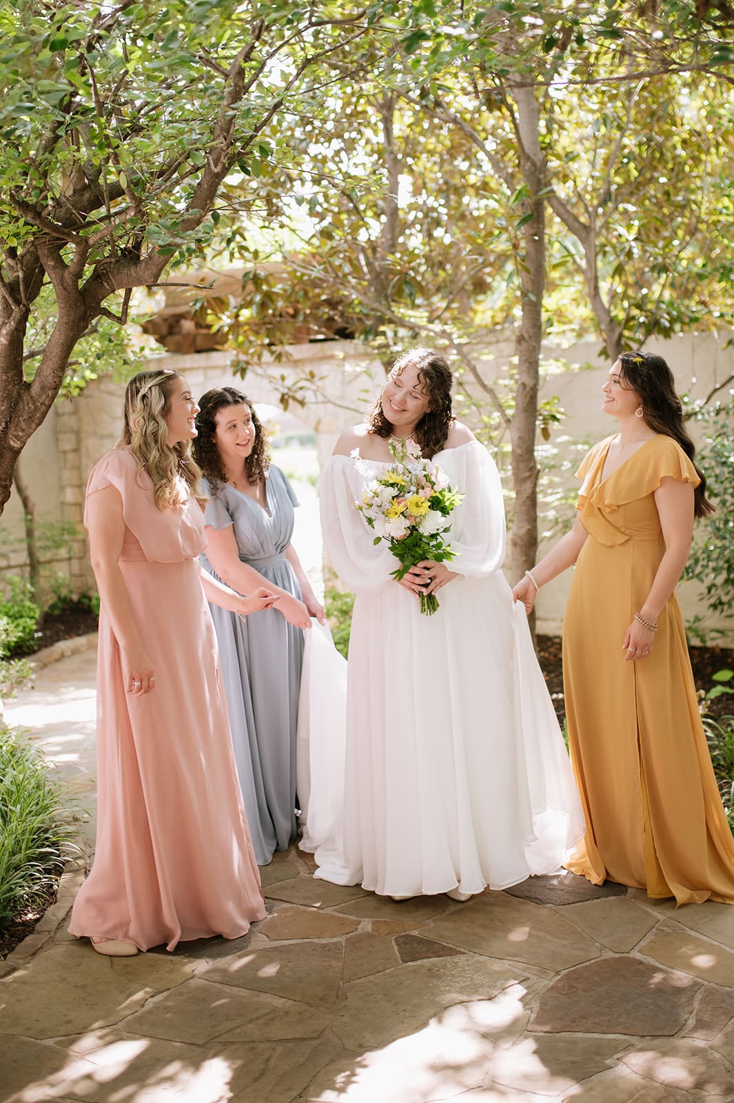Four women in colorful dresses stand outdoors, smiling and laughing. The woman in white holds a bouquet, suggesting she is the bride, while the others hold baskets of flowers.