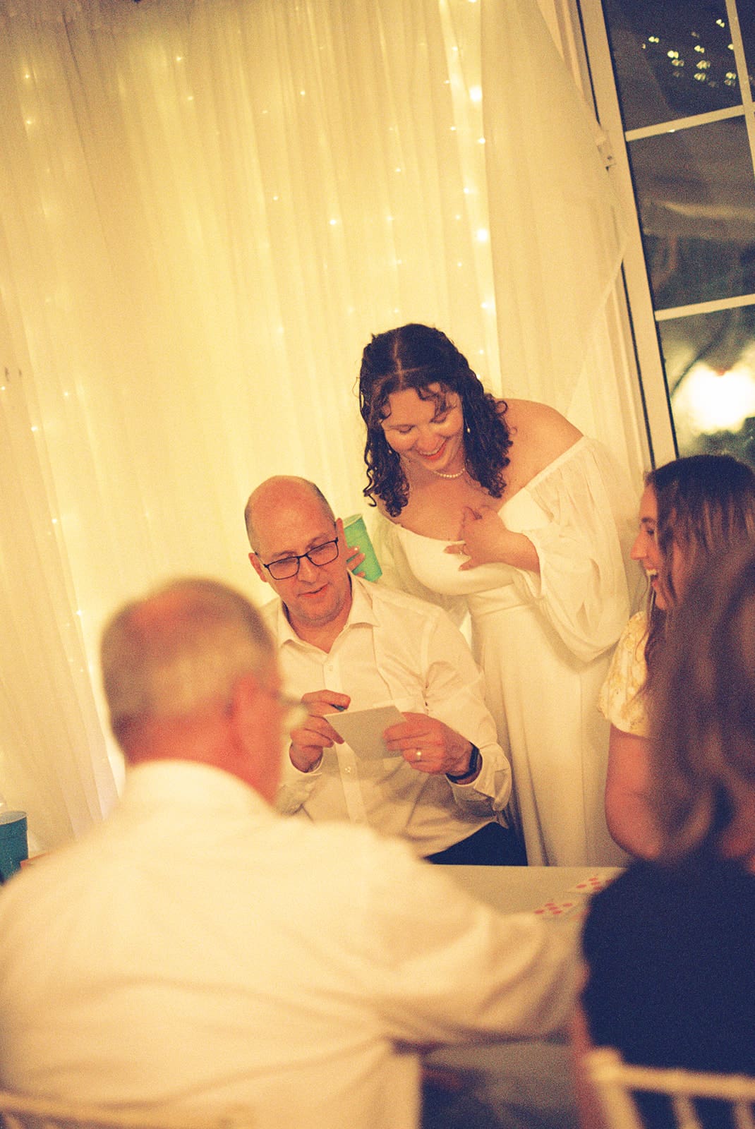 A man reads a card at a table while a woman in a white dress stands beside him, smiling. Two other people are seated nearby. Warm lights decorate the background at Magnolia Terrace in Frisco, Tx