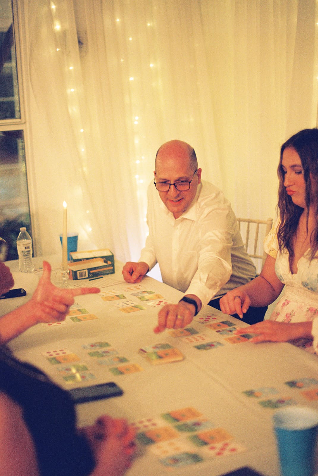 A man reads a card at a table while a woman in a white dress stands beside him, smiling. Two other people are seated nearby. Warm lights decorate the background at Magnolia Terrace in Frisco, Tx