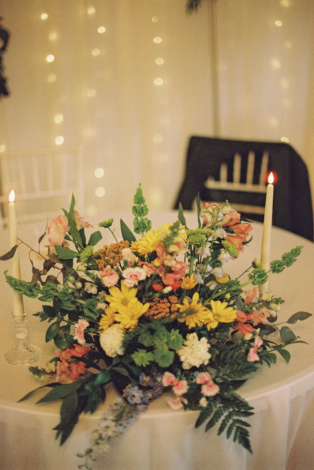 A round table with a large floral arrangement, two lit taper candles, and a white chair draped with a dark jacket in the background.